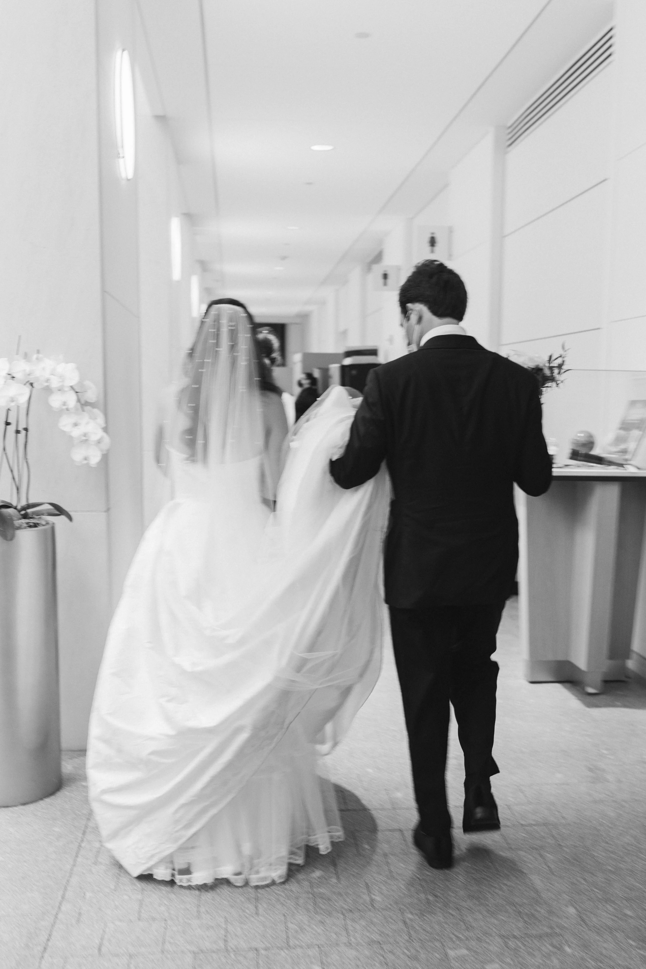 A man in a suit walking behind a bride holding a bouquet in a hospital corridor.