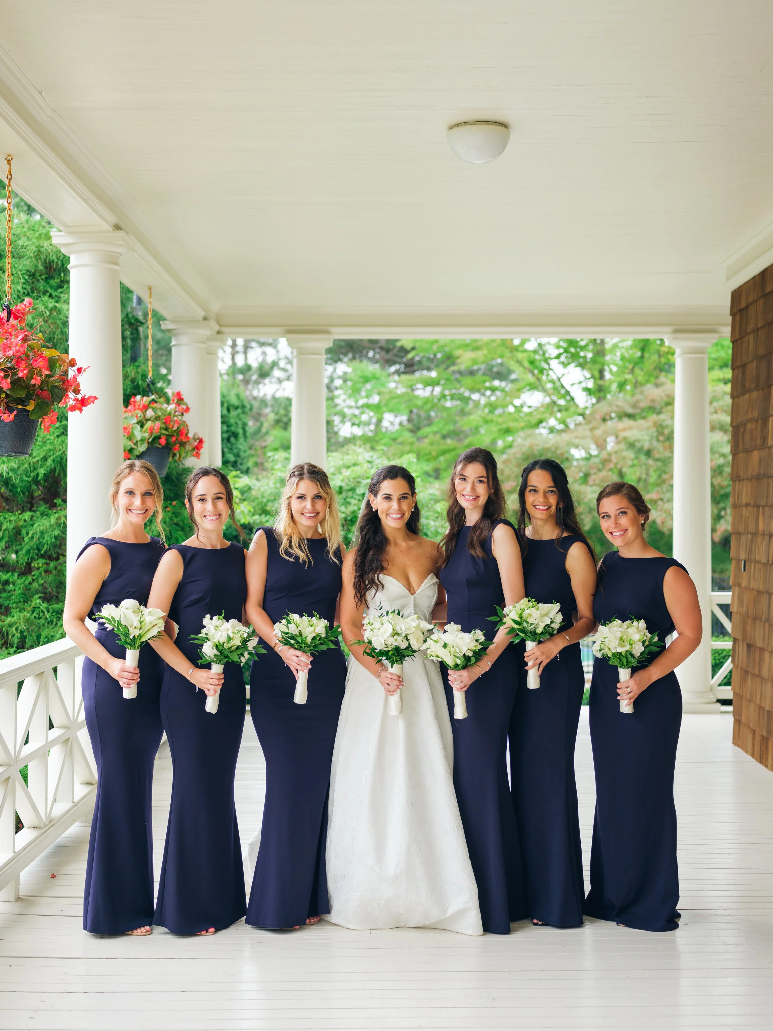Group of seven women in formal dresses, six in navy dresses and one in a white wedding dress, holding bouquets of white flowers, standing on a porch with white columns and greenery in the background.