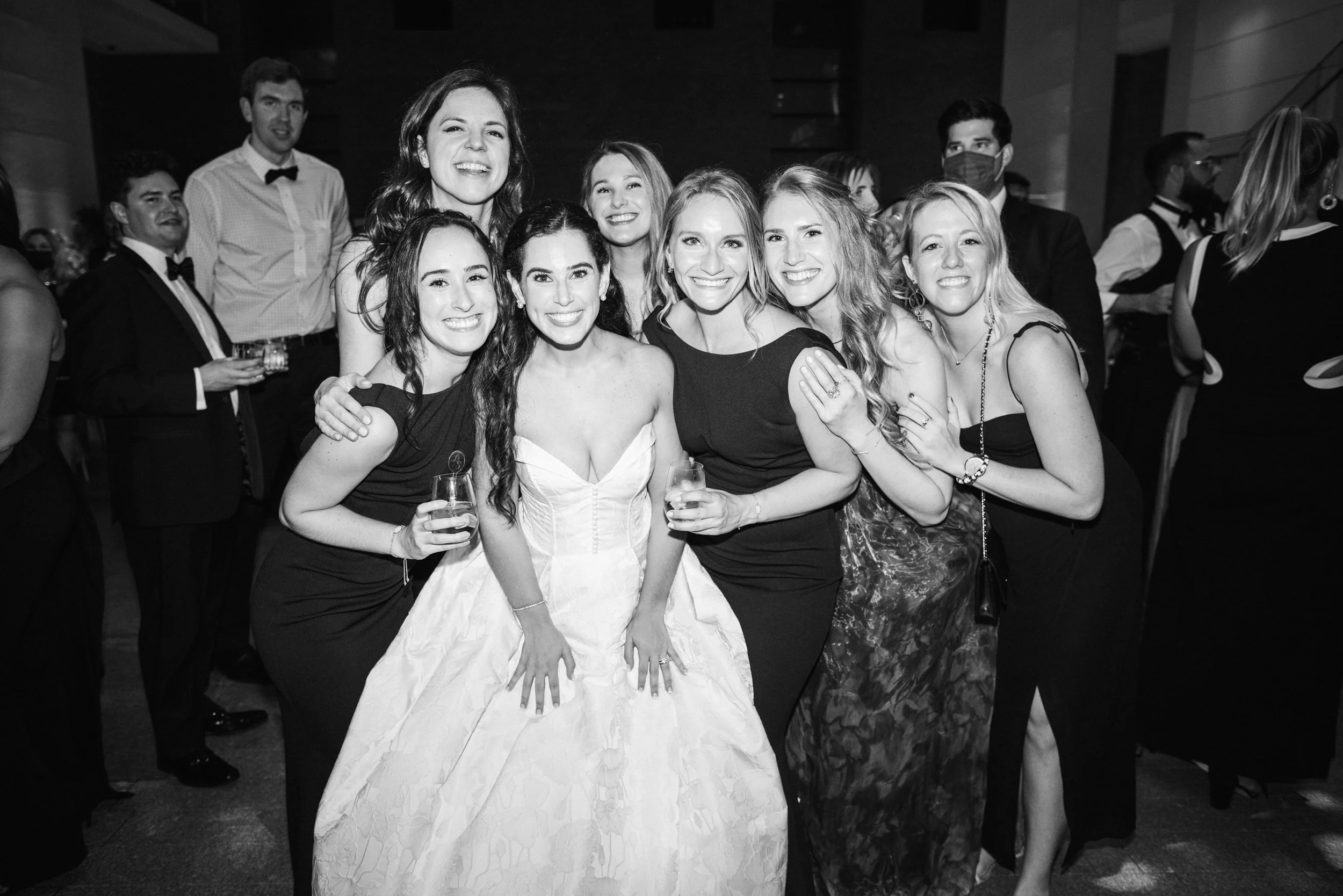 A group of smiling women and men at a formal event, including a woman in a white gown, with others in dresses and tuxedos, some holding drinks, in a dimly lit venue.