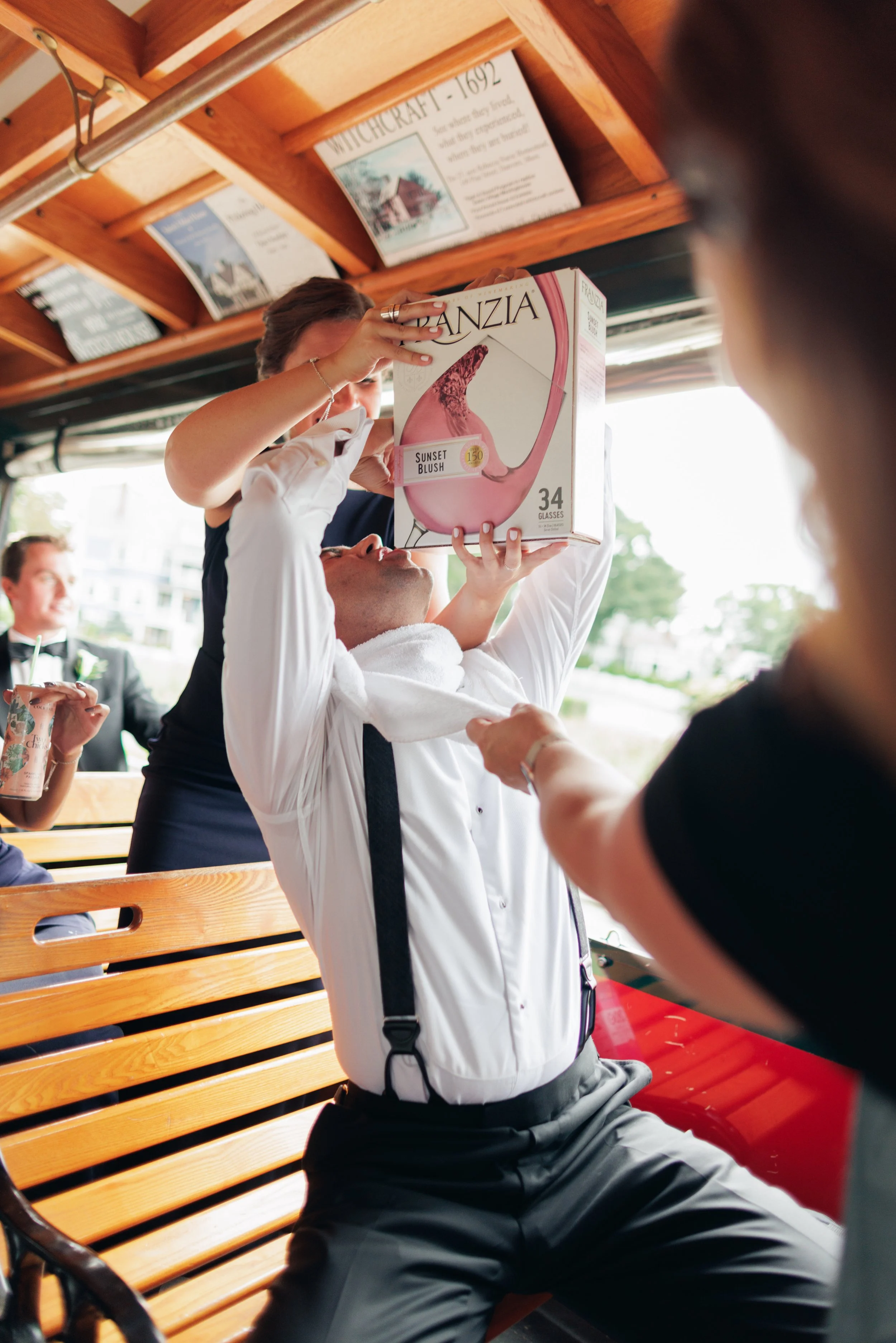 A man in professional attire, wearing a white shirt with suspenders, is sitting on a wooden bench and holding a large box labeled 'Branzia Sunset Blush' over his face. He appears to be passing the box to a woman dressed in a black outfit, in what see