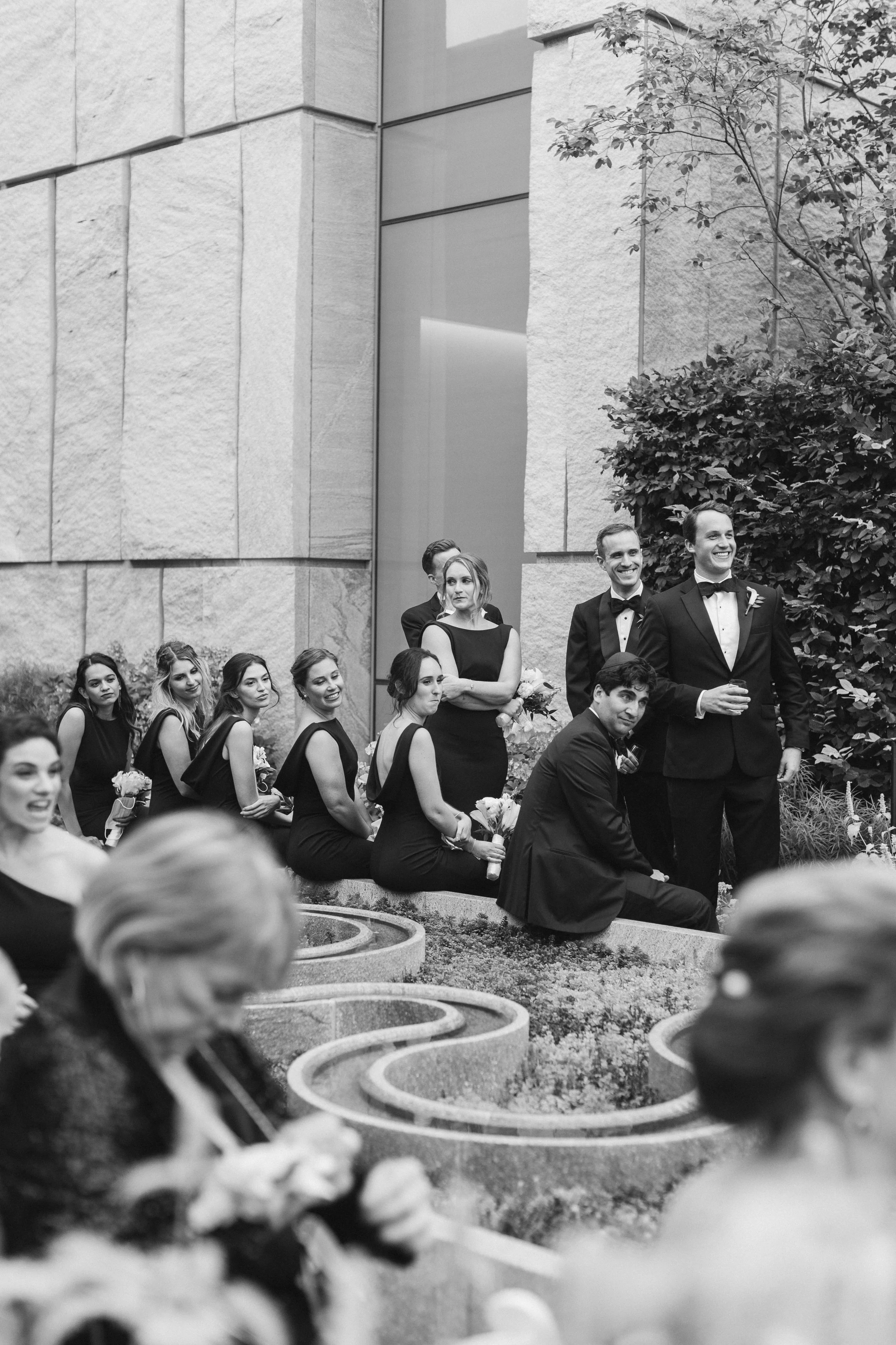 A group of well-dressed people at an outdoor wedding, with men in tuxedos and women in elegant dresses, sitting and standing near a building with a textured stone wall and bushes.