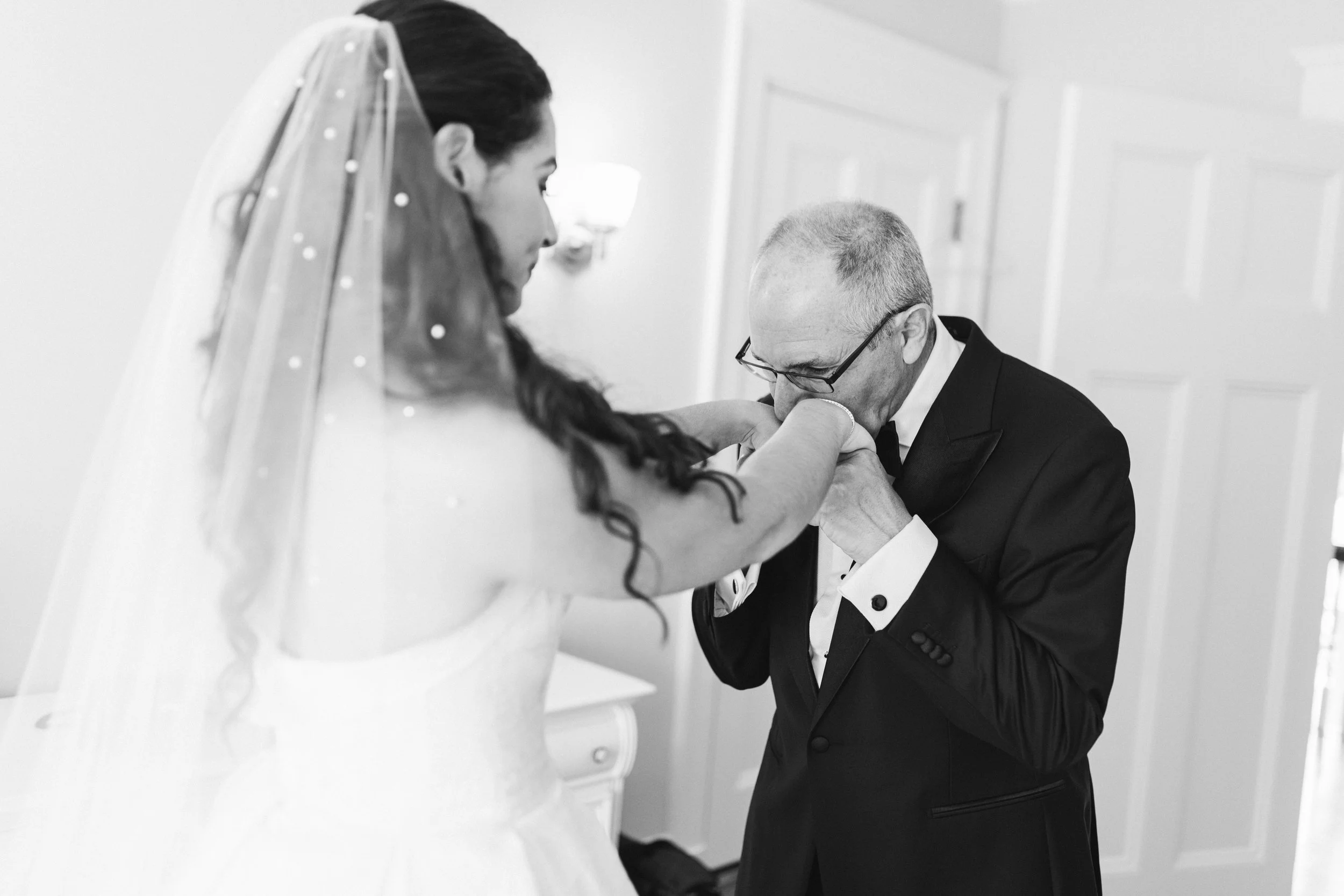A groom dressed in a tuxedo is kneeling and kissing the hand of a bride, who is wearing a veil adorned with small embellishments, in an indoor setting with a plain wall and door in the background.