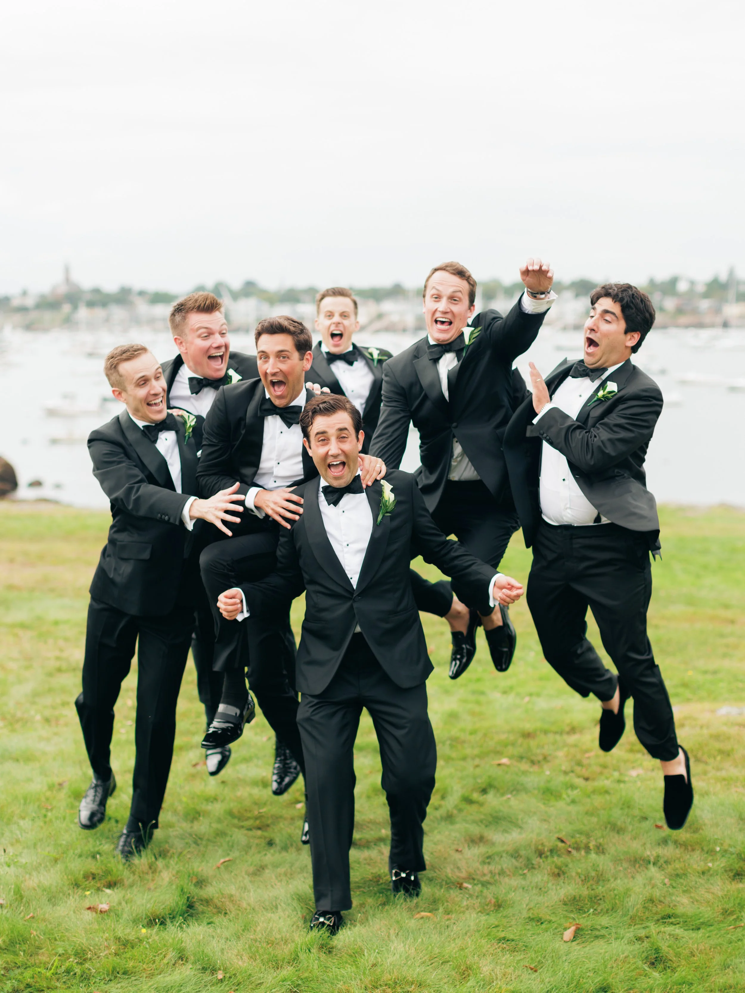 A group of seven men in tuxedos jumping and celebrating on a grassy area, with a marina and boats in the background.
