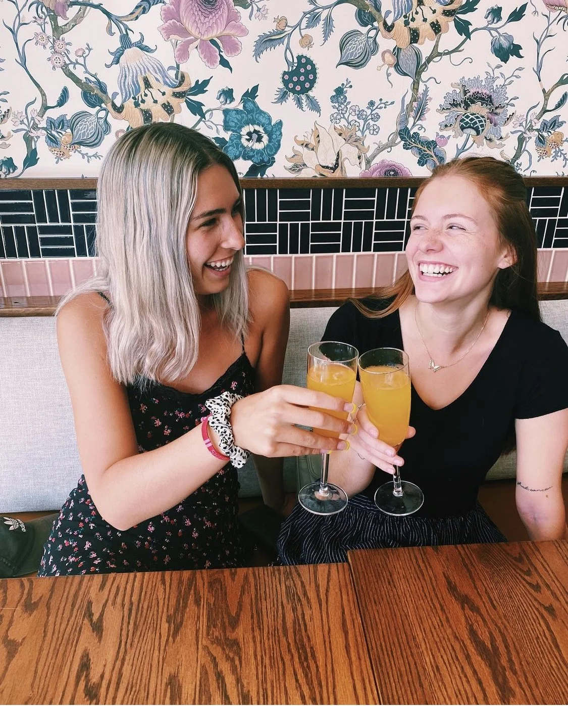 Two young women sitting at a wooden table, smiling, and holding glasses of orange-colored drinks, toasting in a restaurant with floral wallpaper and a patterned black and pink tile wall behind them.