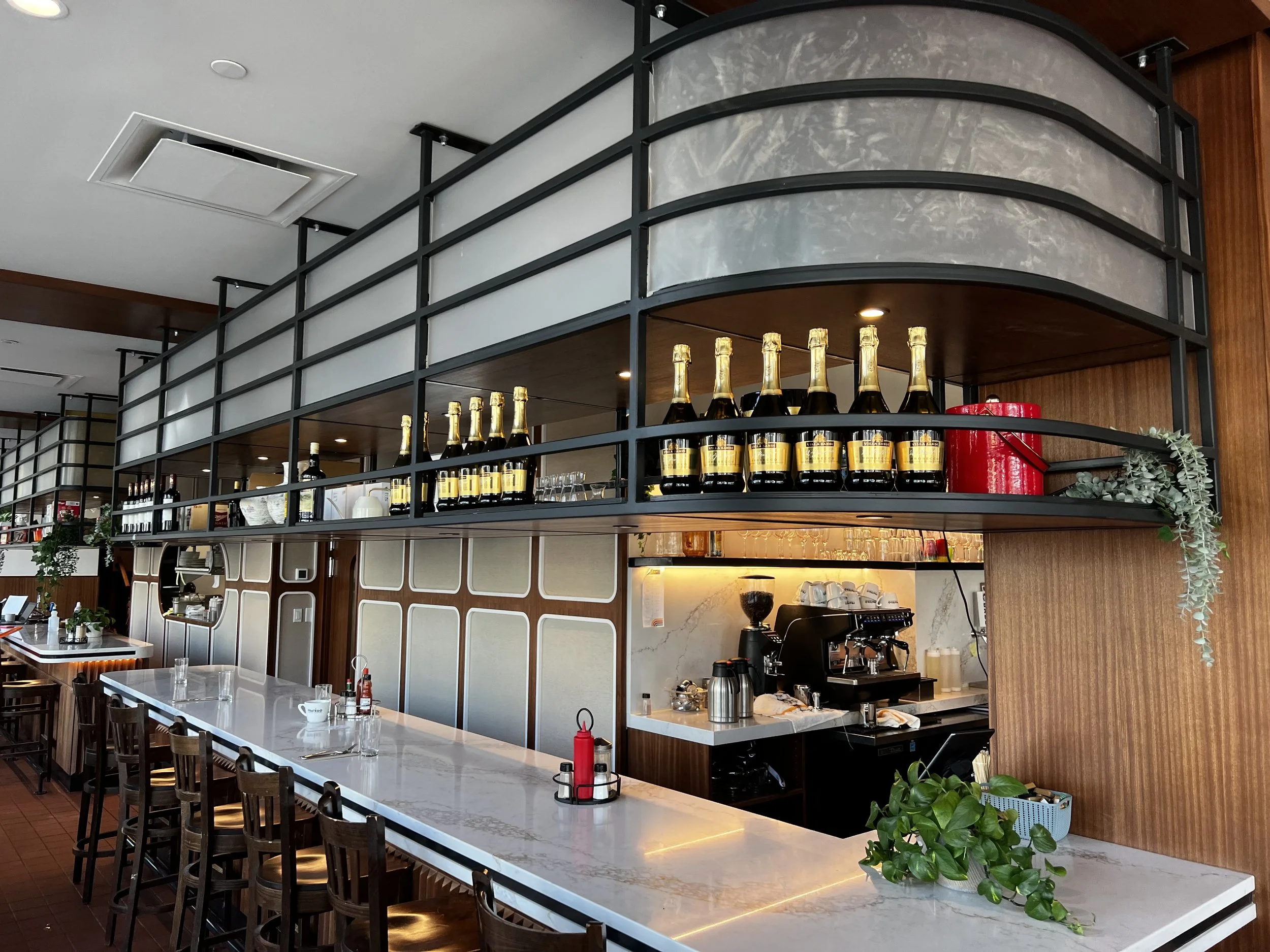 Bar area with a row of champagne bottles on a curved black metal shelf above a white marble counter; counter has a plant, condiments, and glasses; behind the bar, there is a coffee machine, cups, and various equipment, with warm lighting and wood-paneled walls.