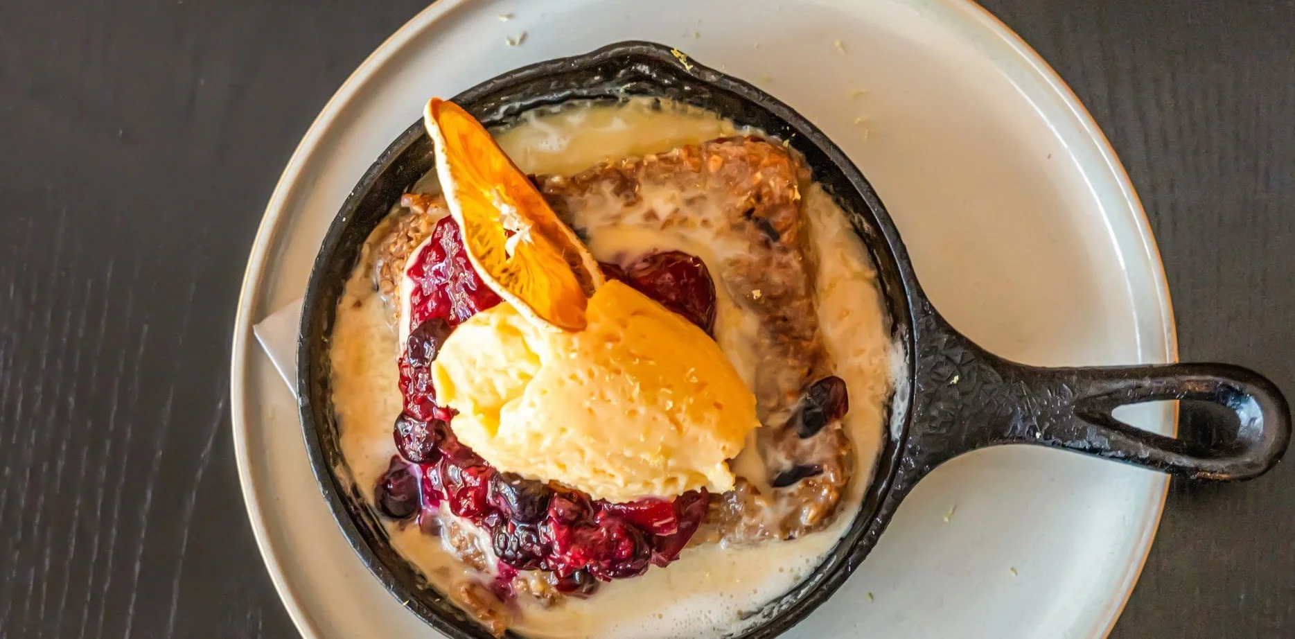 A dessert served in a small cast iron skillet with a scoop of vanilla ice cream, fresh cranberries, orange slices, and a creamy sauce, set on a white plate on a dark table.