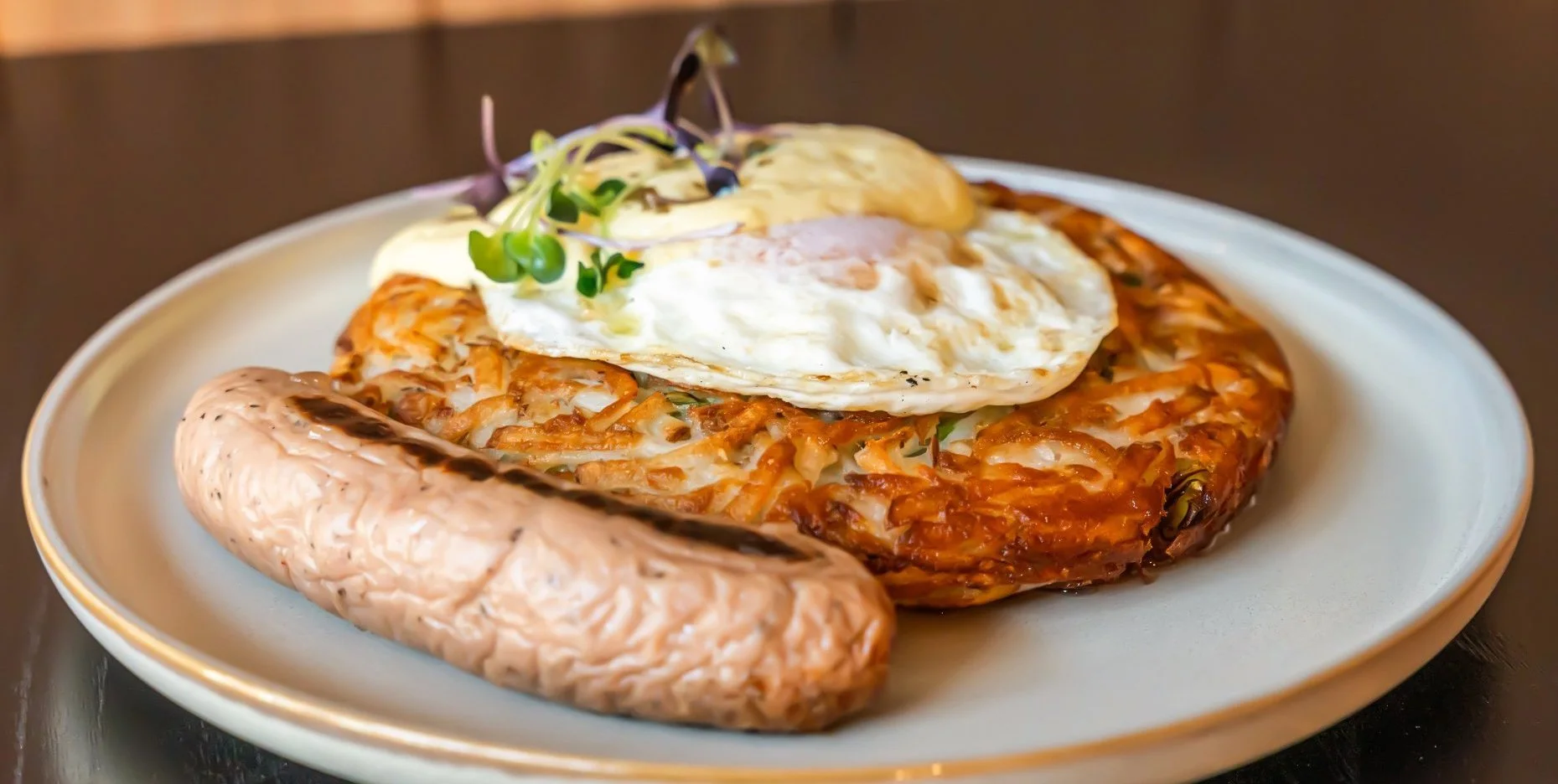 Plate with fried egg on top of kimchi pancake, side of sausage, garnished with microgreens.