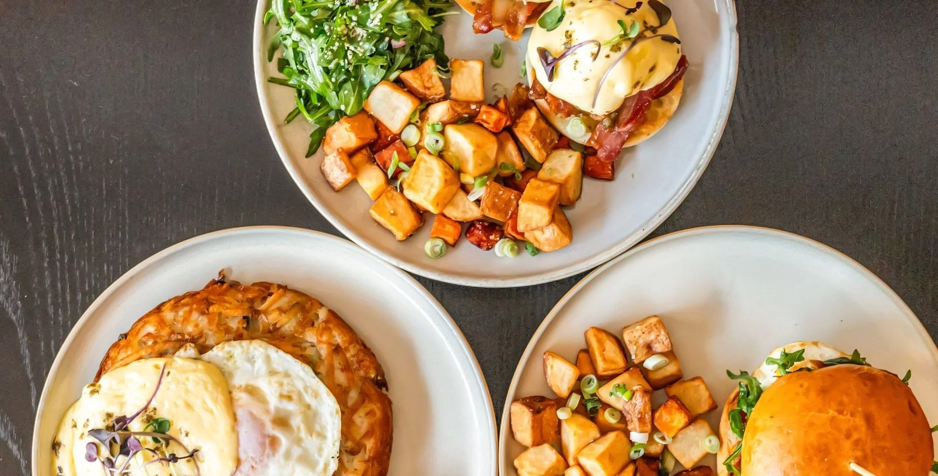 Three plates of brunch dishes on a dark table: one with a fried egg on top of a hash with vegetables, another with roasted potatoes, greens, and an egg, and the third with a sandwich bun and greens.