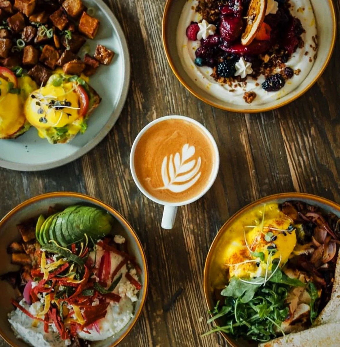 A top-down view of a wooden table with five dishes and a cup of coffee. The dishes include scrambled eggs with avocado and salsa on toast, a bowl of berry granola with yogurt and berries, sliced avocado on rice with vegetables, a breakfast dish with 