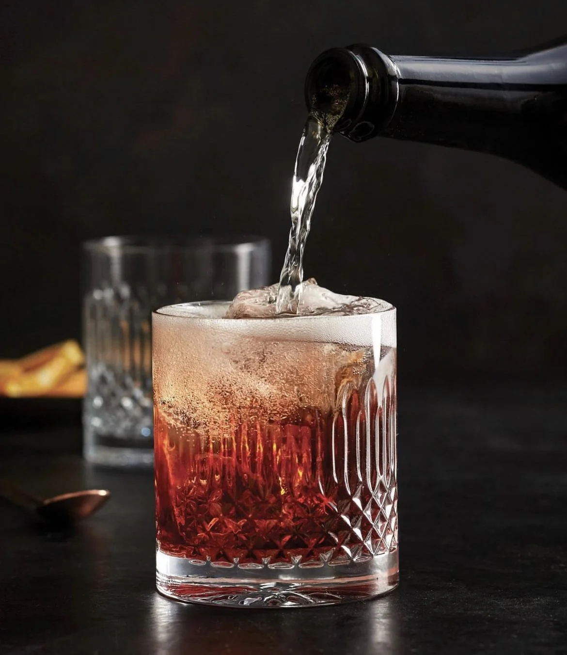 A glass with a textured pattern is being filled with a dark, fizzy liquid from a bottle against a dark background.