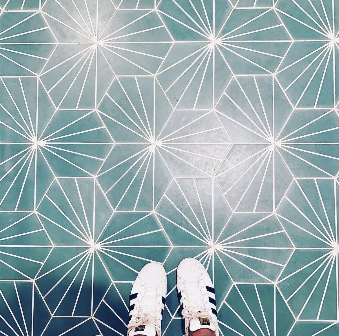 View of feet wearing white Adidas sneakers with black stripes standing on a patterned teal and white tile floor.