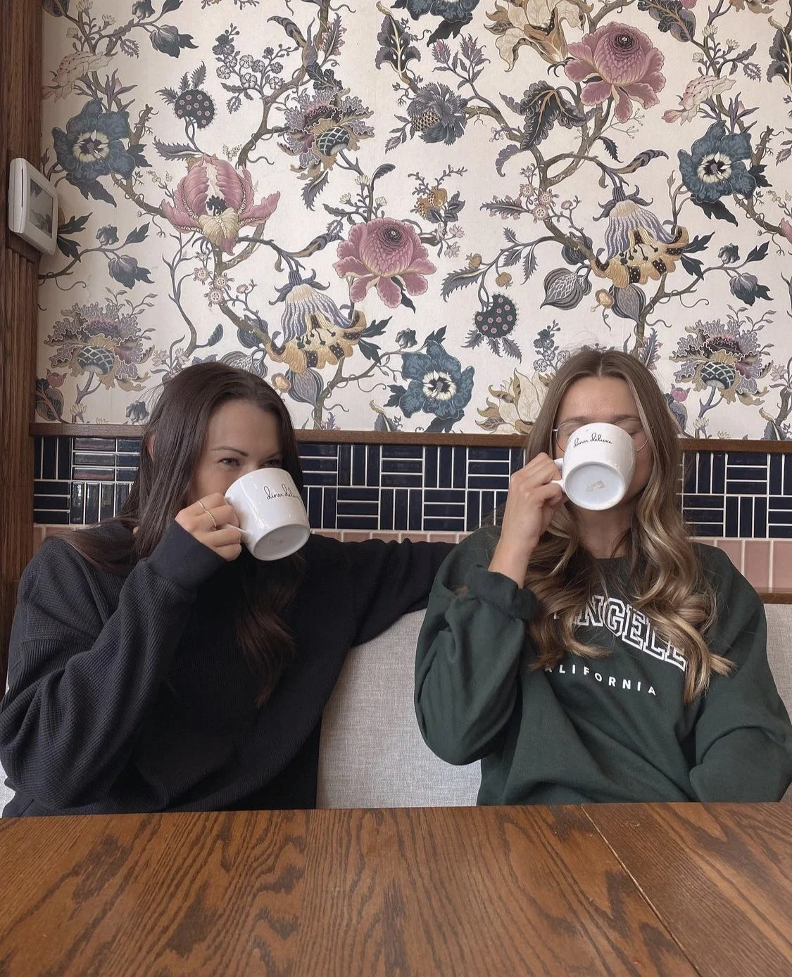 Two women sitting at a wooden table, drinking from white mugs, with a floral patterned wallpaper background.