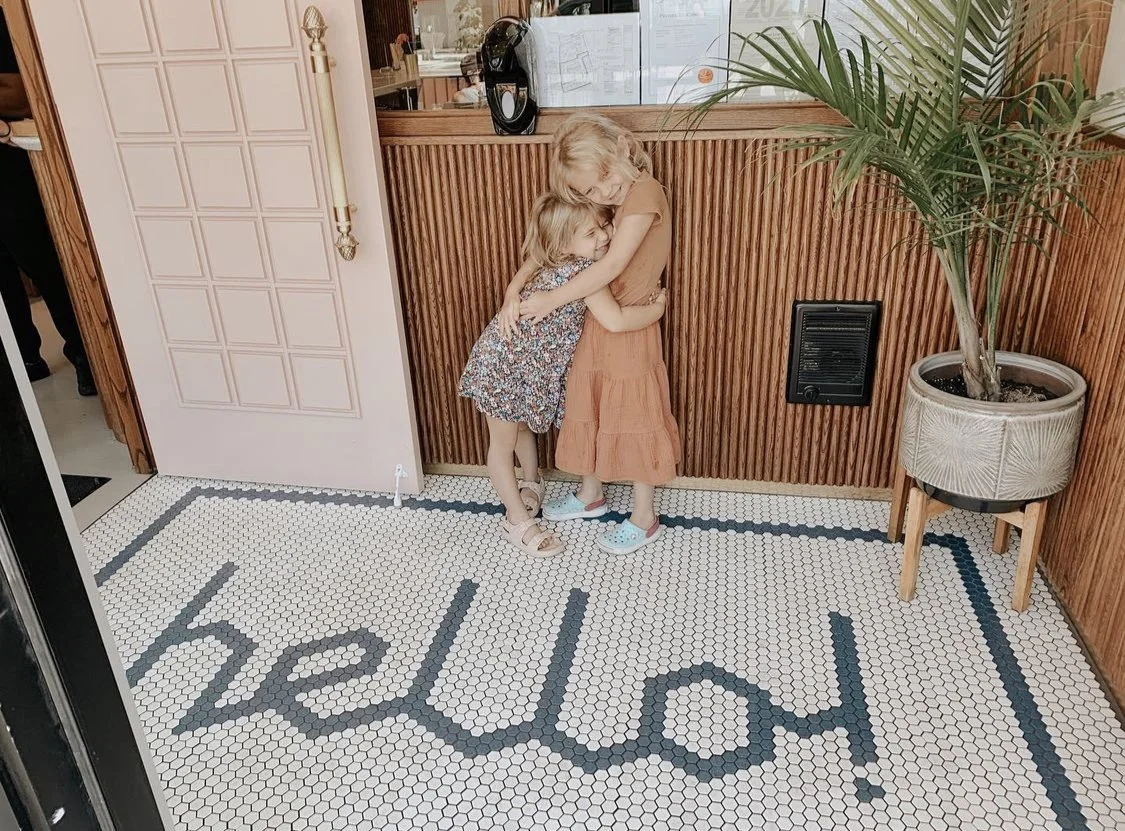 Two young girls hugging on a tiled floor with the word 'hello' spelled out in black tiles. One girl has blonde hair and is wearing a floral dress, the other has light brown hair and is wearing an orange dress. They are next to a wooden panel wall with a potted plant on a stand.