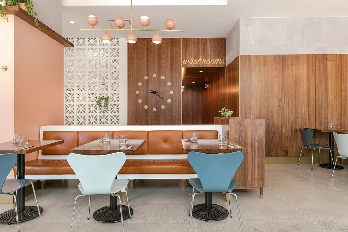 Interior of a modern restaurant with wooden and pastel pink walls, a large clock, a sign that reads 'washrooms', and various tables and chairs.