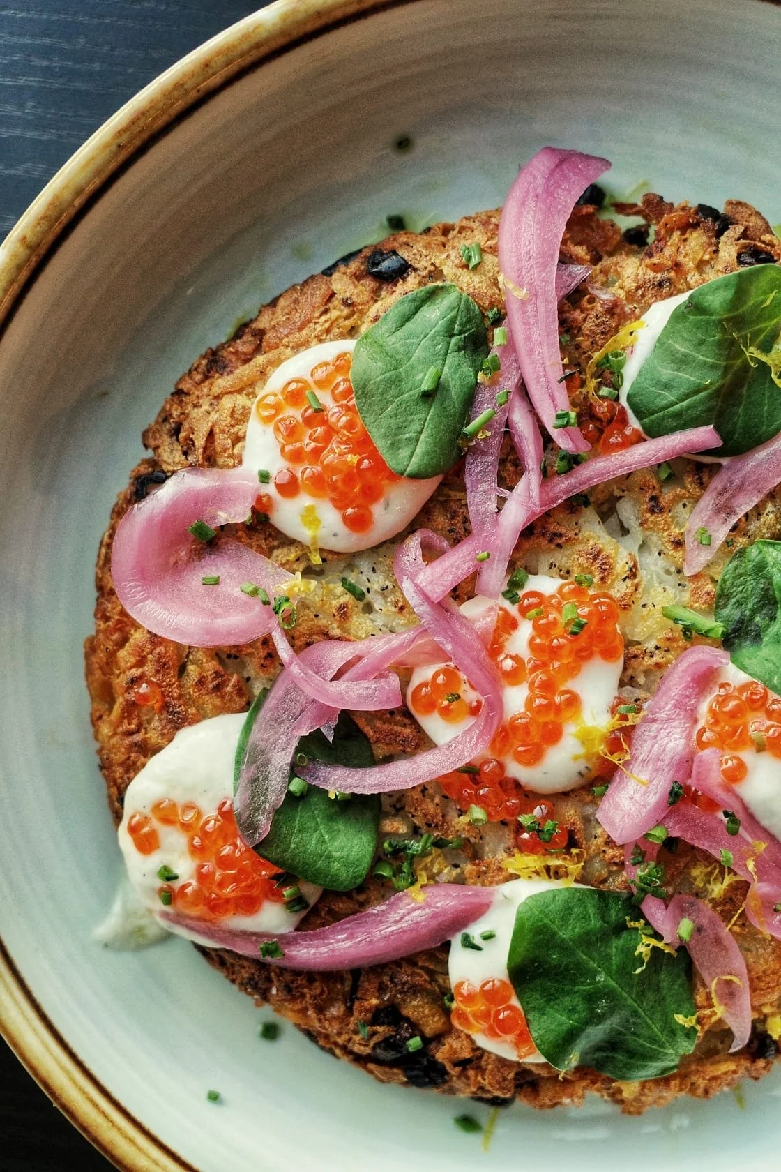 Close-up of a toasted bread slice topped with whipped sour cream, orange salmon roe, thin slices of pickled red onions, fresh basil leaves, chopped chives, and lemon zest.