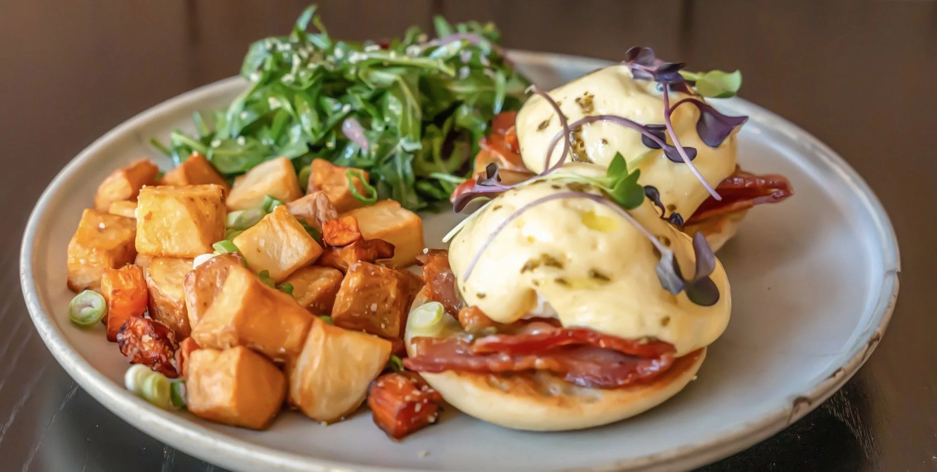 Plate of poached eggs Benedict with microgreens, roasted sweet potatoes, and a side of greens.