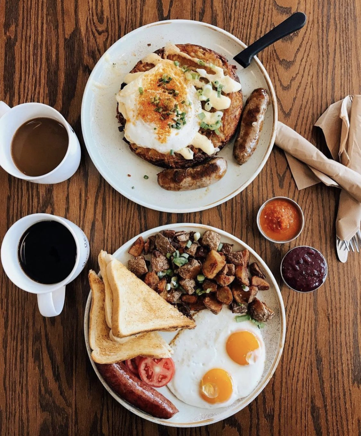 Two breakfast plates on a wooden table. Top plate has eggs benedict with sausage links, and the bottom plate has two sunny-side-up eggs, toast, sausage, potatoes, and tomato slices, along with cups of coffee and small bowls of salsa and jam.