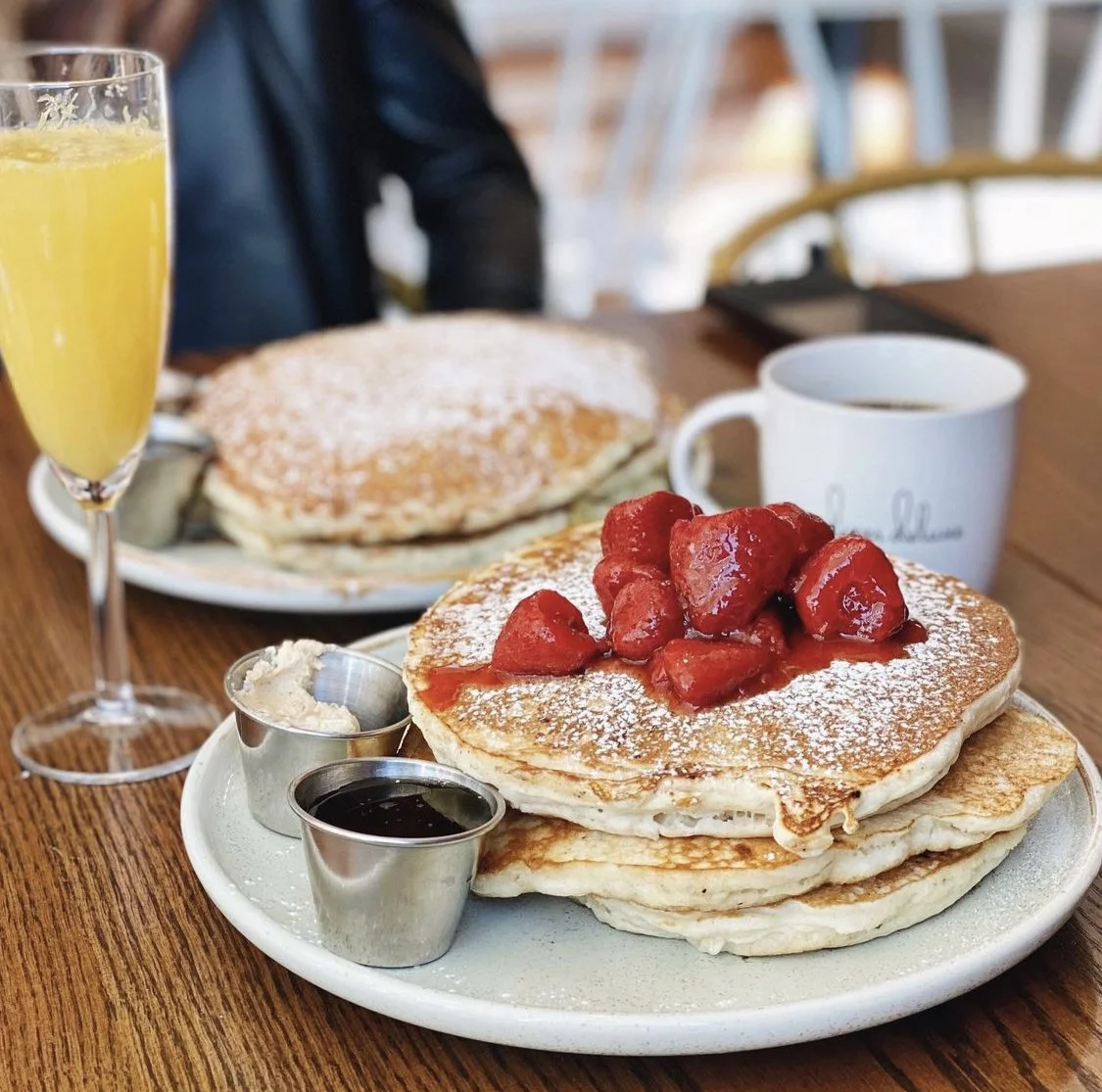 A breakfast plate with three pancakes topped with strawberries and powdered sugar, served with syrup and butter. Accompanied by a cup of coffee, a glass of orange juice, and a second plate with more pancakes in the background.