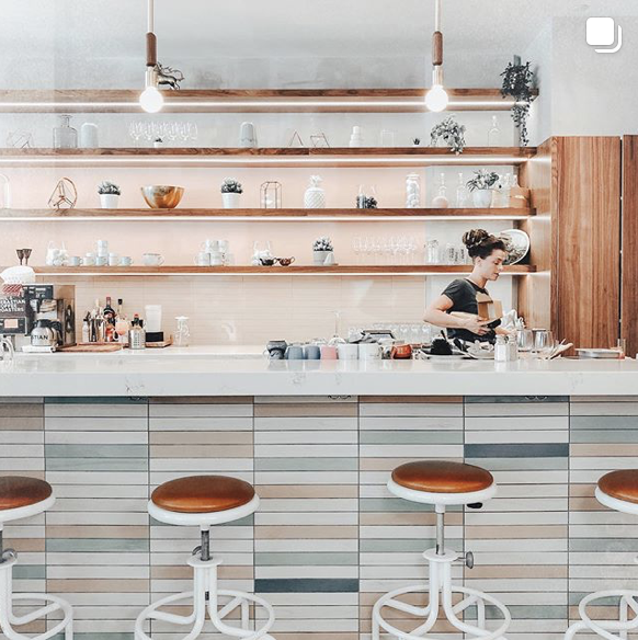 A woman preparing coffee behind a bar in a modern, minimalistic café with shelves of glassware and potted plants.