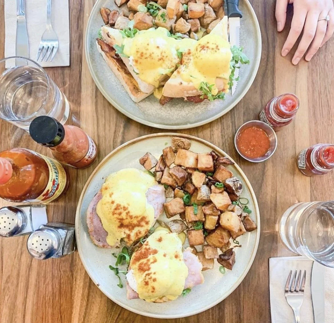 Two plates of eggs Benedict with ham, plus sautéed potatoes on a wooden table, surrounded by condiments, glasses of water, and utensils.