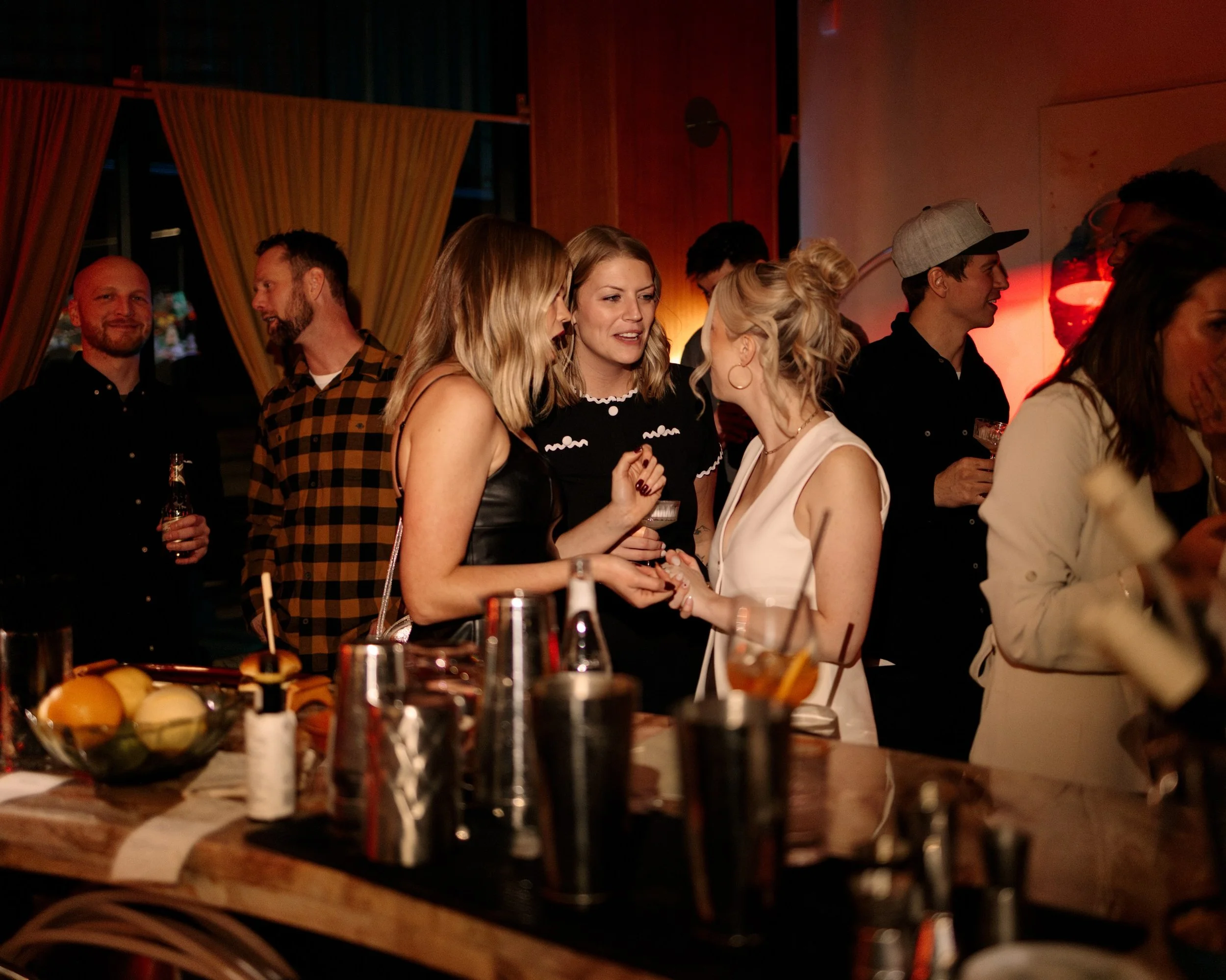 People socializing at a bar or party, with women engaged in conversation near the countertop, drinks and snacks visible, in a dimly lit environment.