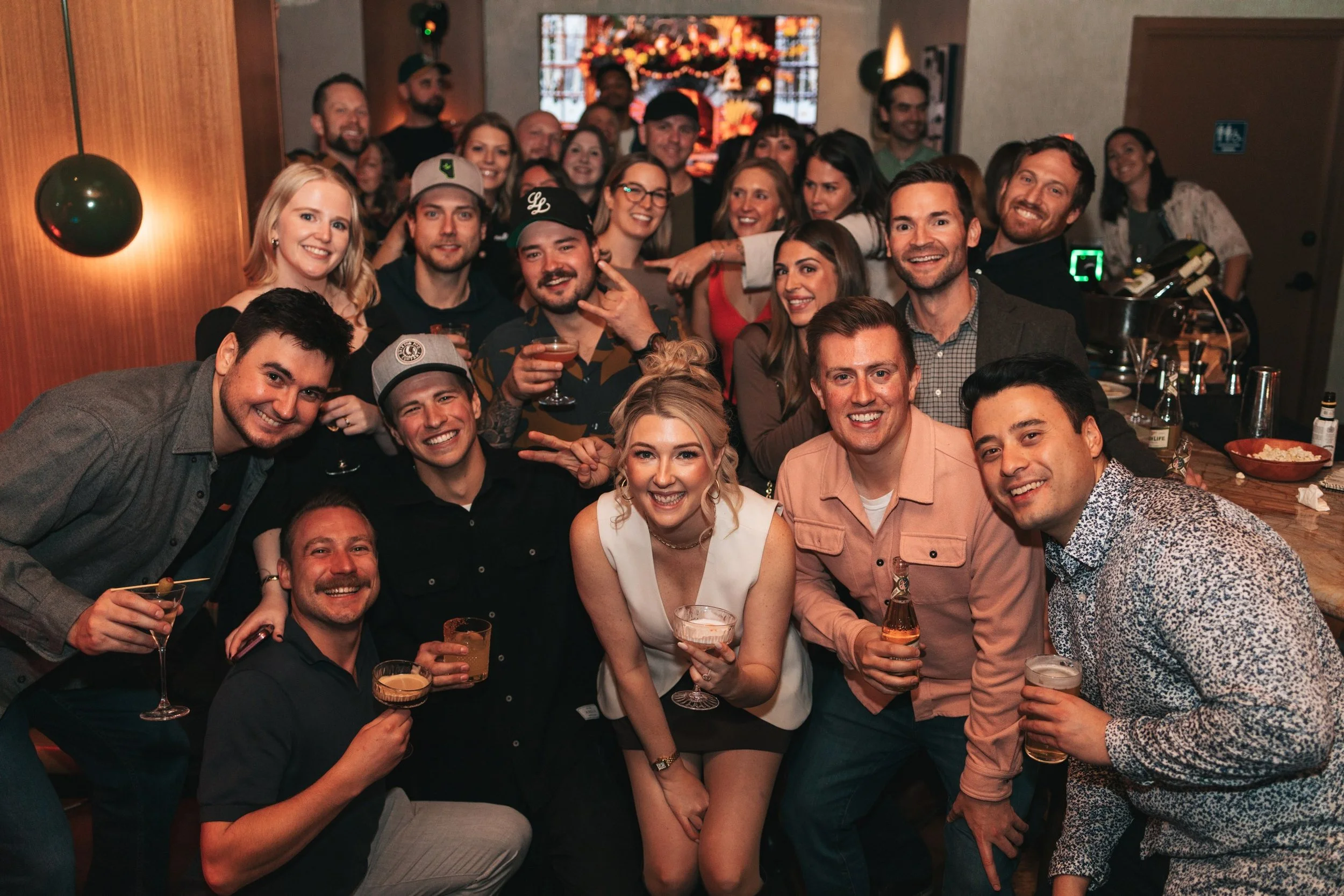 A large group of people gathered at a celebration or party, smiling at the camera, with some holding drinks. The setting appears to be indoors, decorated for a festive occasion.