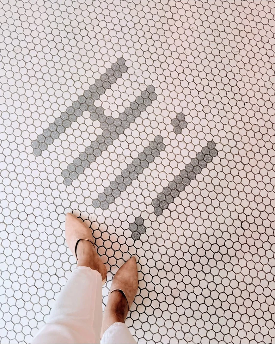 A person wearing tan high-heeled shoes and white pants standing on a white hexagonal tiled floor with three parallel lines of small dark gray tiles.