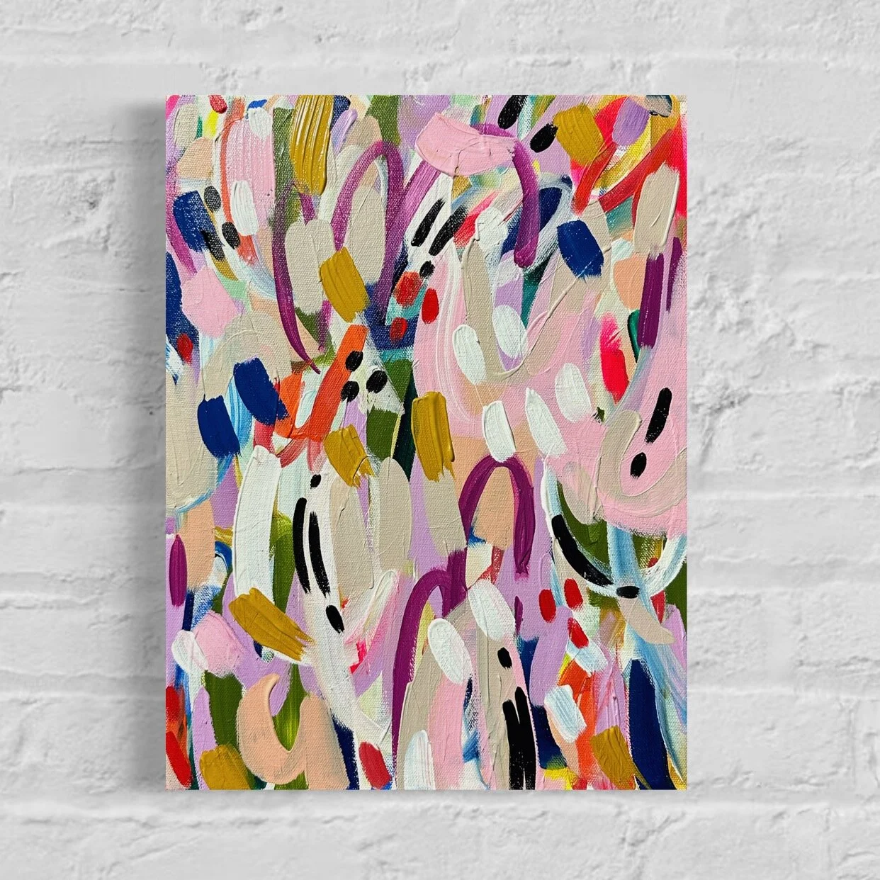 Abstract colorful painting with brushstrokes of pink, white, purple, blue, red, black, and mustard on canvas, hung on a white brick wall.