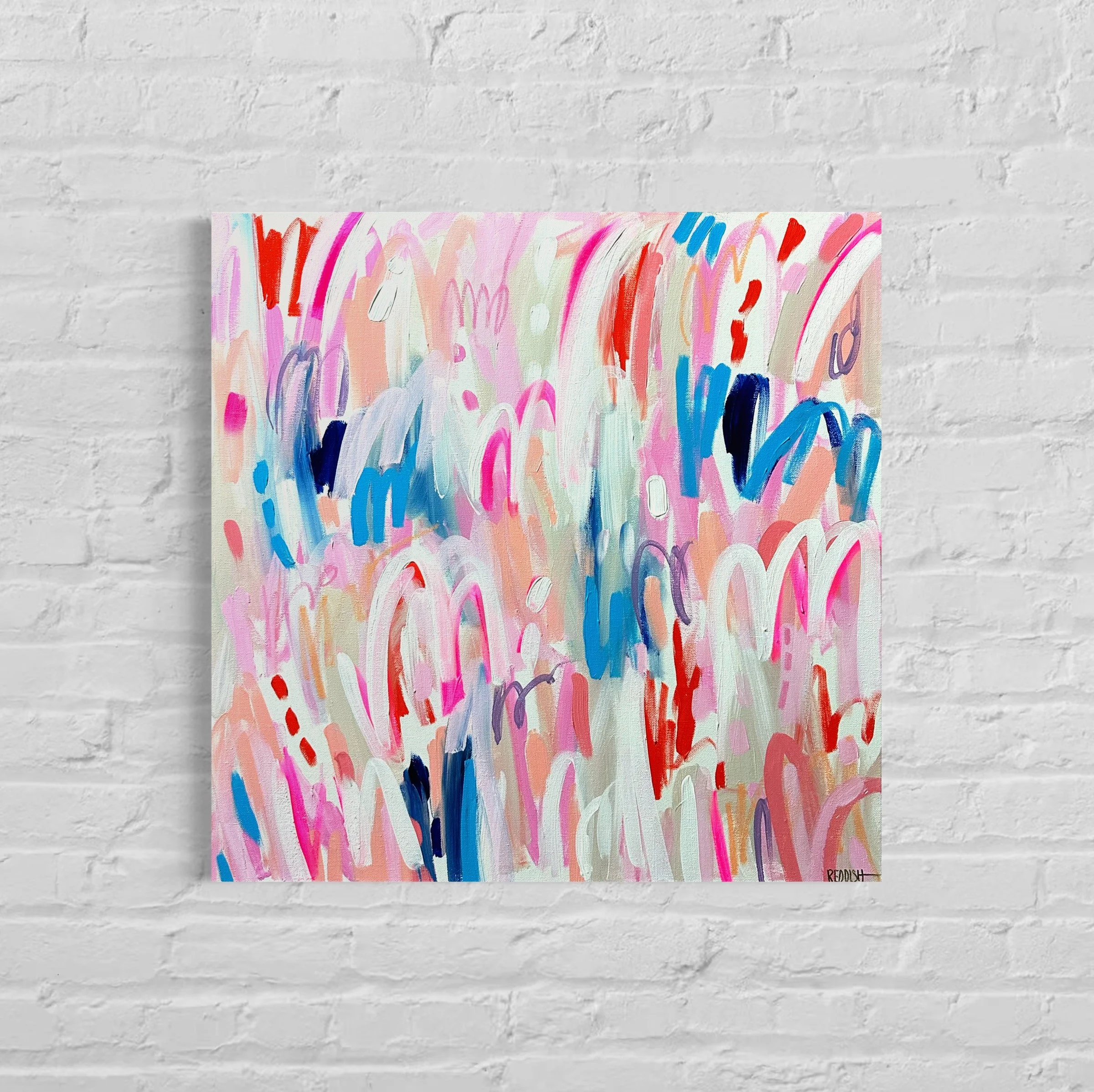 Abstract colorful expressionist painting on canvas with brushstrokes in pink, red, blue, orange, purple, and white, hanging on a white brick wall.