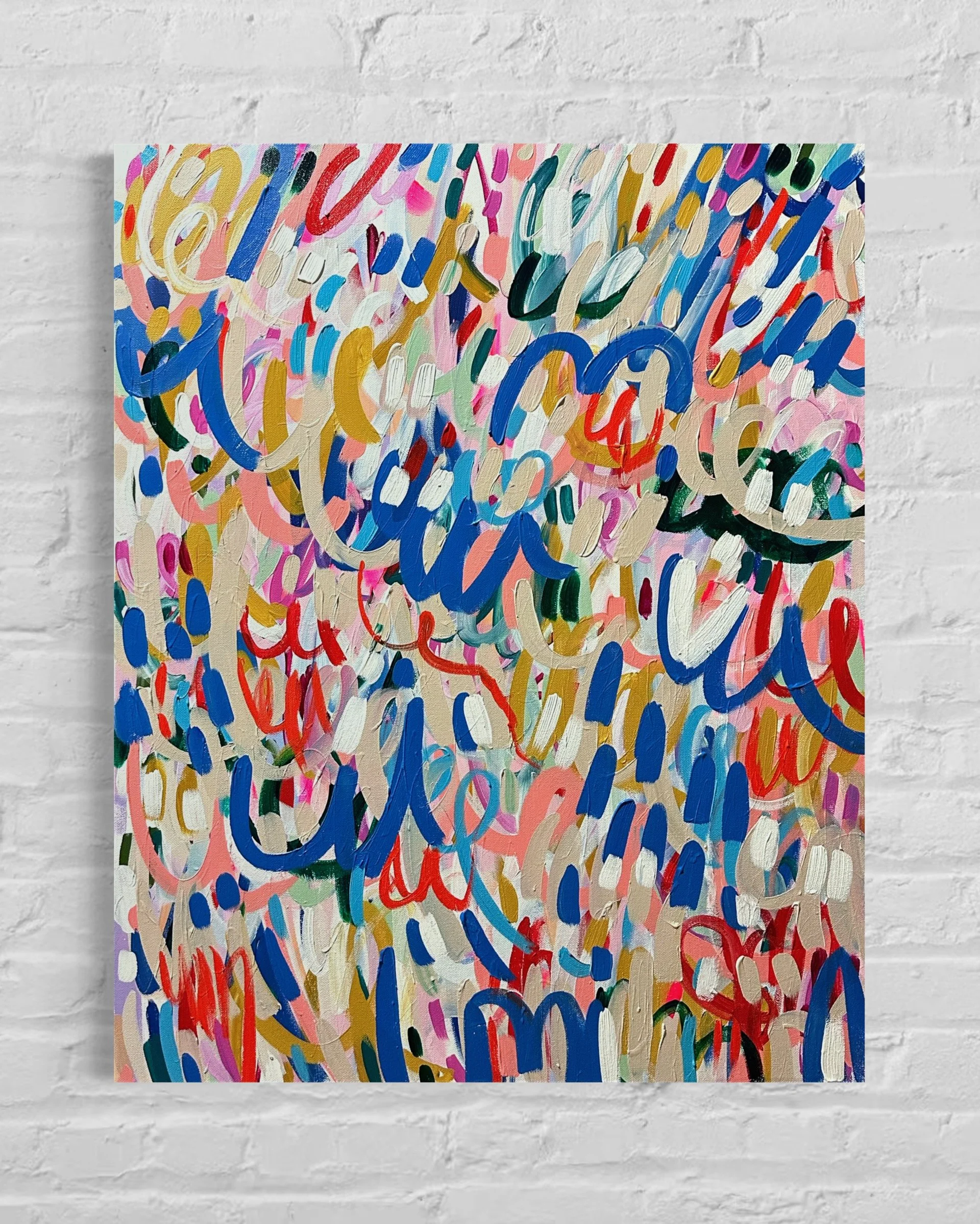 Abstract colorful painting with various swirls and brushstrokes in blue, red, pink, green, black, white, and gold, on a white canvas hung against a white brick wall.