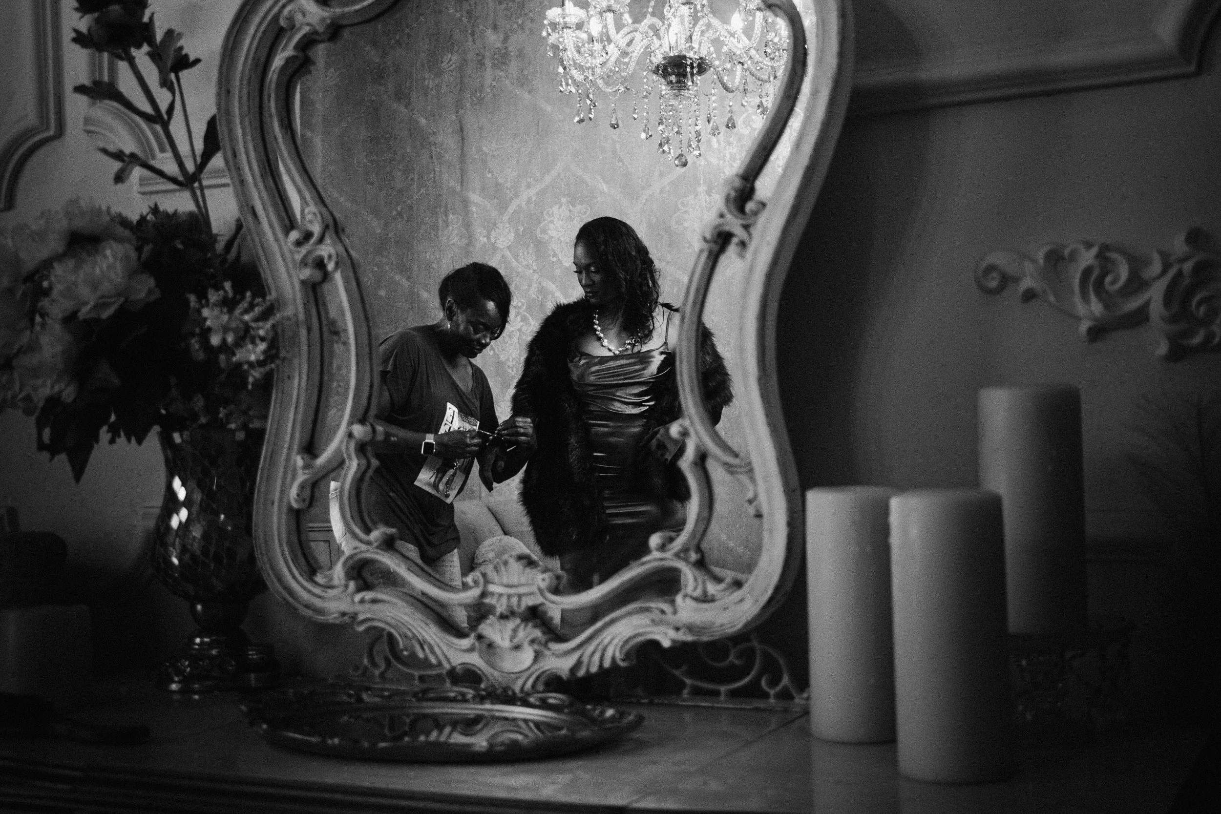 A black and white photo of two women looking at something in a mirror that has an ornate frame. The room has a chandelier hanging from the ceiling and floral wallpaper. One woman is wearing a dress with a strapless top and a necklace, while the other woman is wearing a top with a jacket.