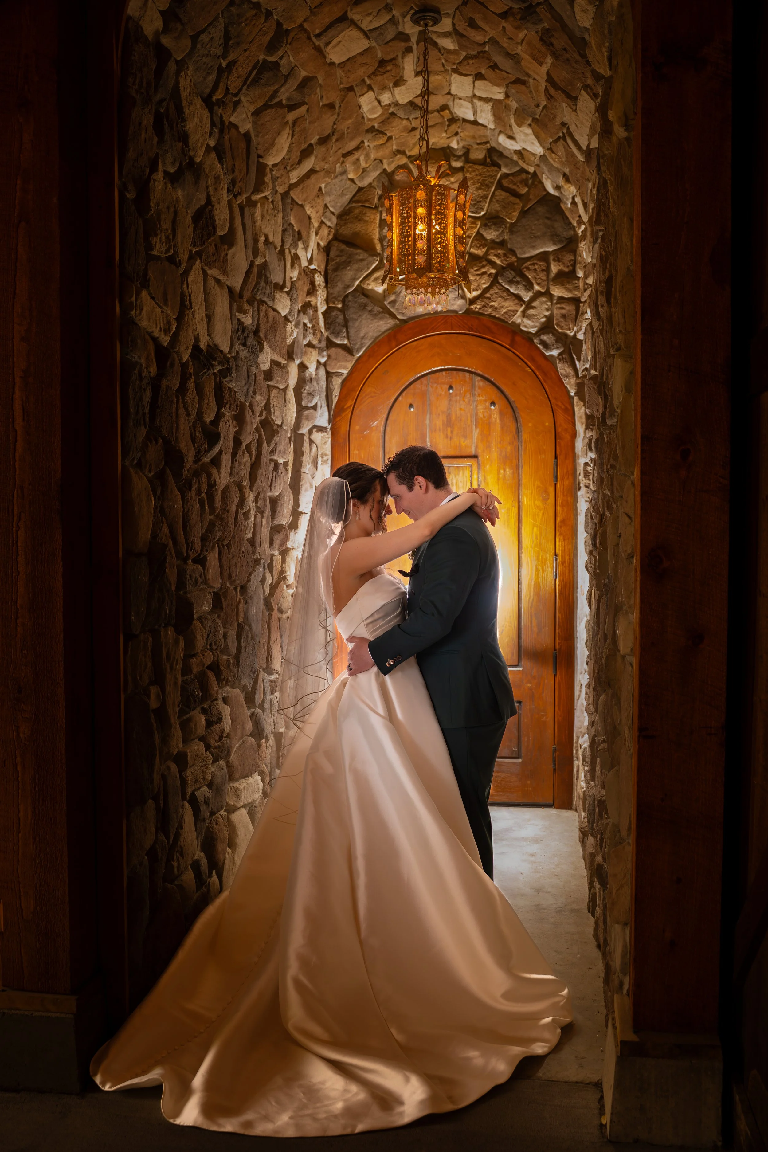 A bride and groom share a dance in a rustic stone hallway illuminated by a hanging chandelier, with a wooden door at the end of the corridor.