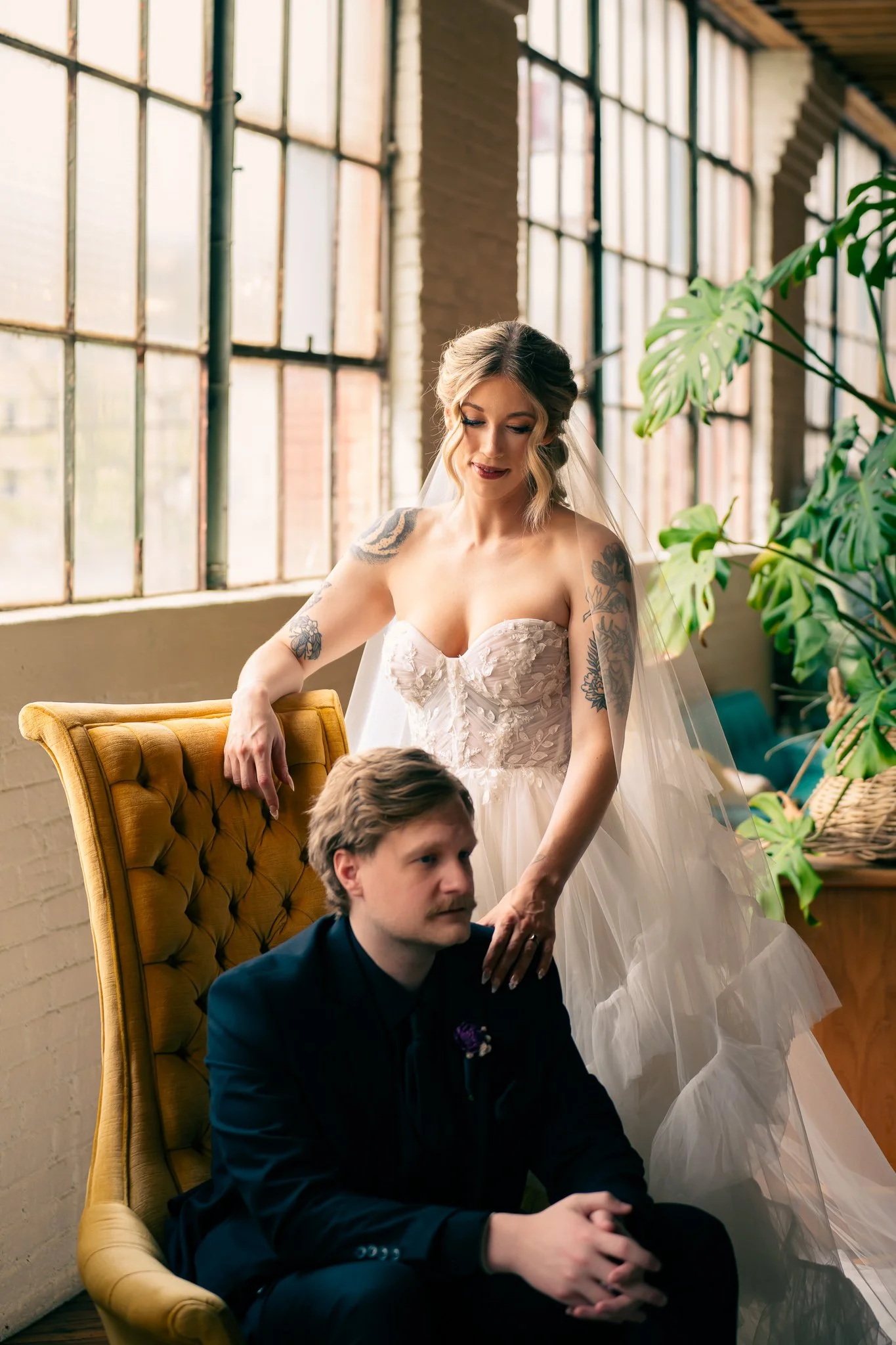 A bride in a white wedding dress with tattoos on her arms, standing behind a seated groom in a black suit, in a room with large industrial-style windows and greenery.
