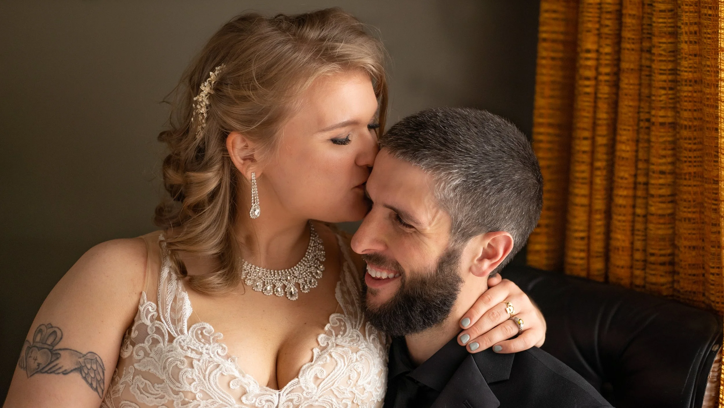 A woman with blonde hair and tattoos on her arm, wearing jewelry and a lace dress, kisses a man with short hair and a beard, who is smiling with his eyes closed.