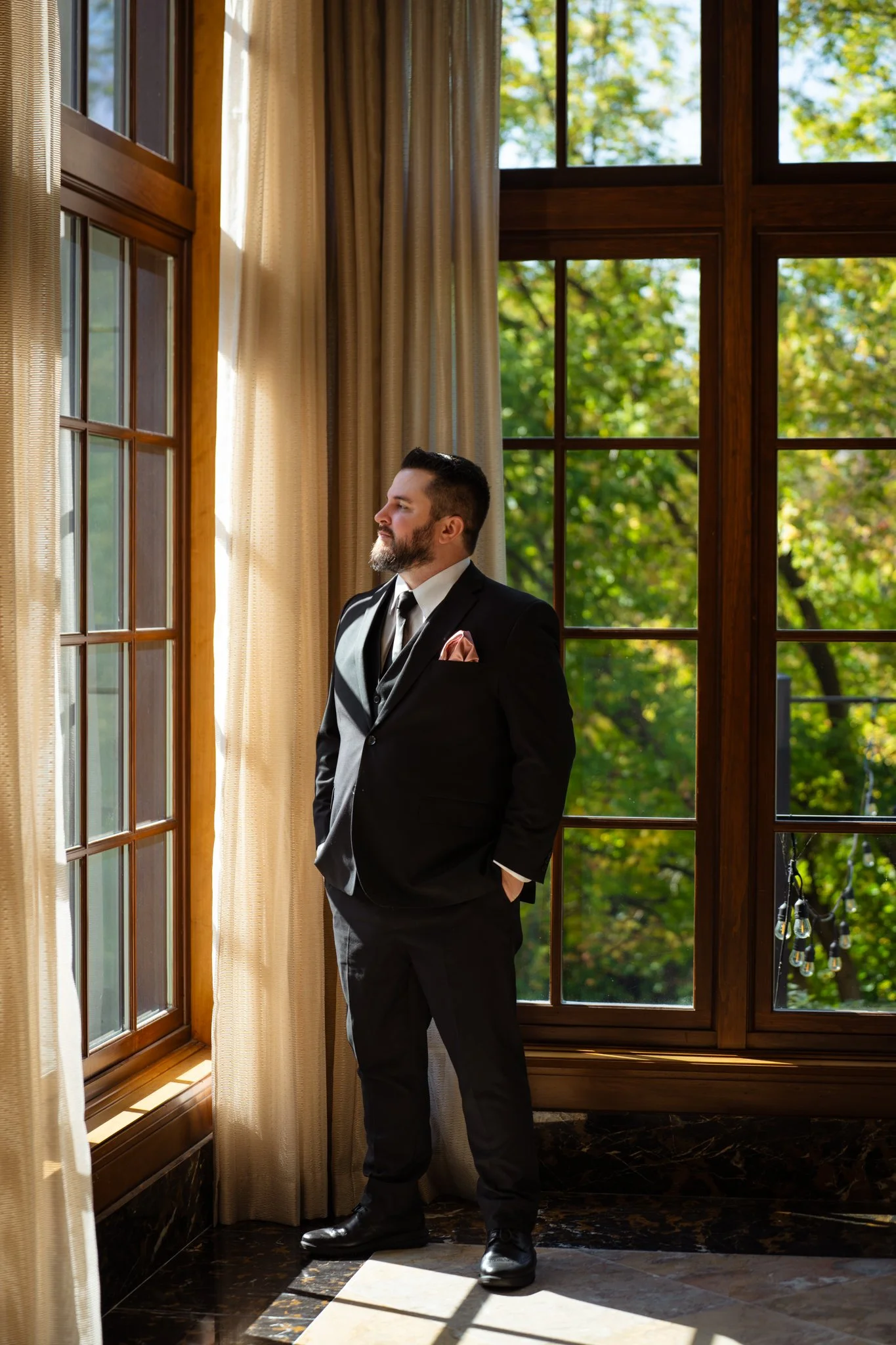 A man in a black suit with a pink pocket square stands in front of a large window with beige curtains, looking outside on a sunny day. Trees with green leaves are visible outside.