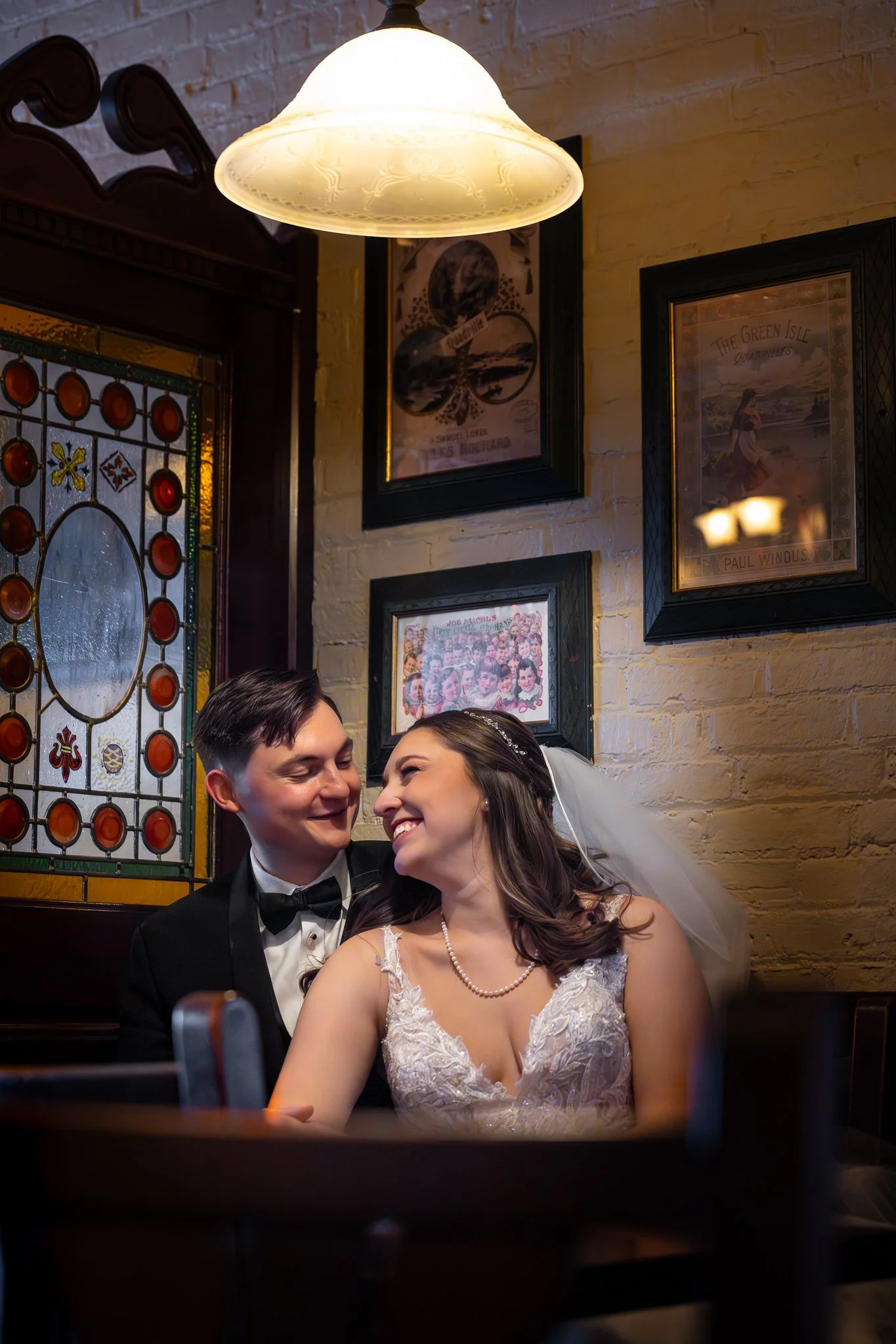 A newlywed couple sitting close together in a restaurant, smiling and looking into each other's eyes, with colorful vintage-style artwork on the wall behind them and warm lighting overhead.