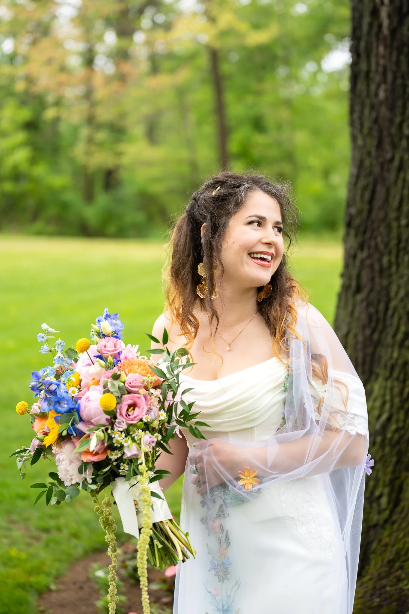 A smiling woman in a white dress with a sheer floral overlay is holding a large colorful bouquet of flowers. She is standing outdoors near a tree in a lush green forest setting.