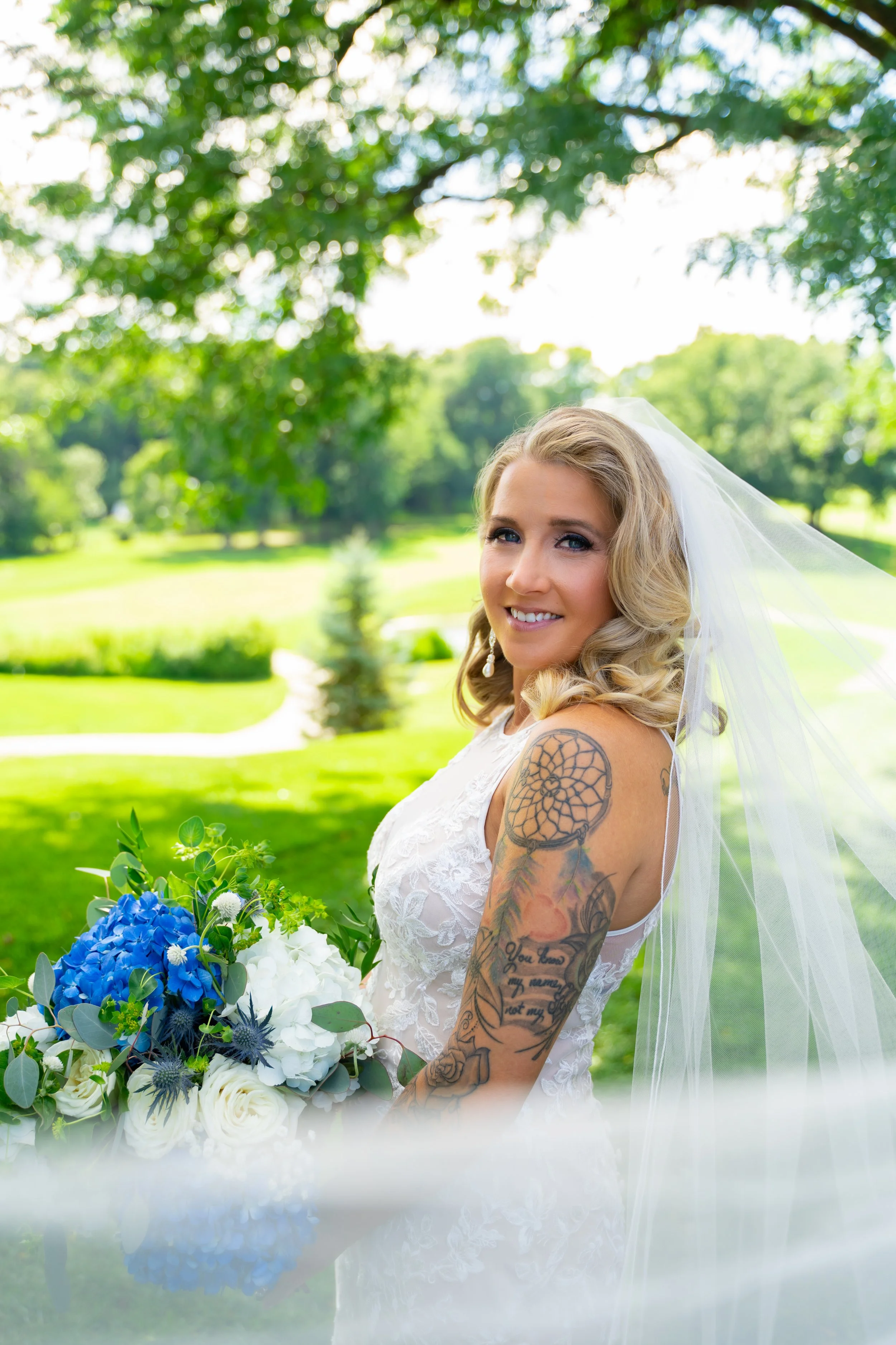 A smiling bride with blonde hair in loose curls holding a bouquet of white and blue flowers for wedding photography in Michigan.
