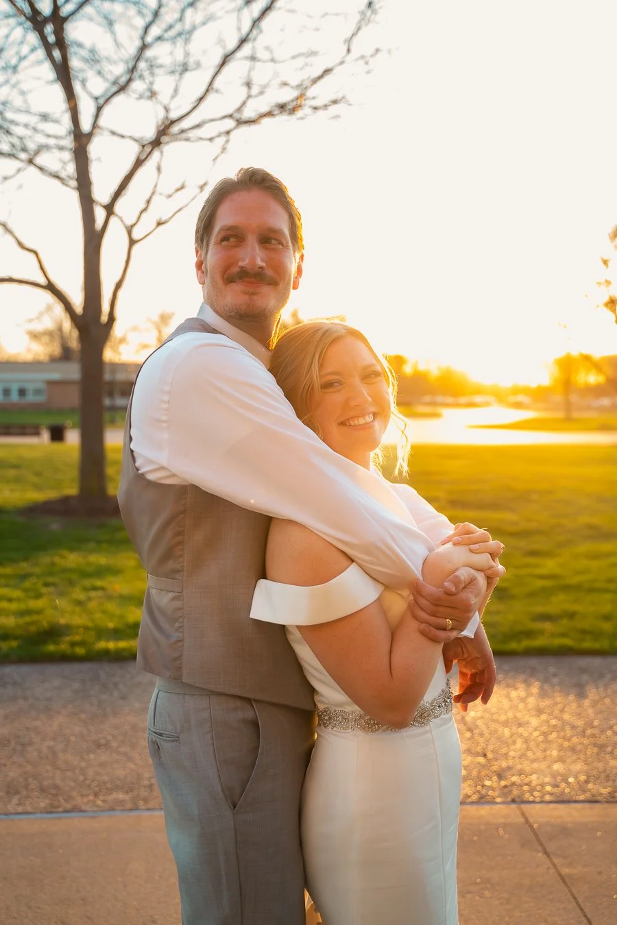 A smiling couple in wedding attire hugging outdoors at sunset in a park captured by a wedding photographer in Michigan.