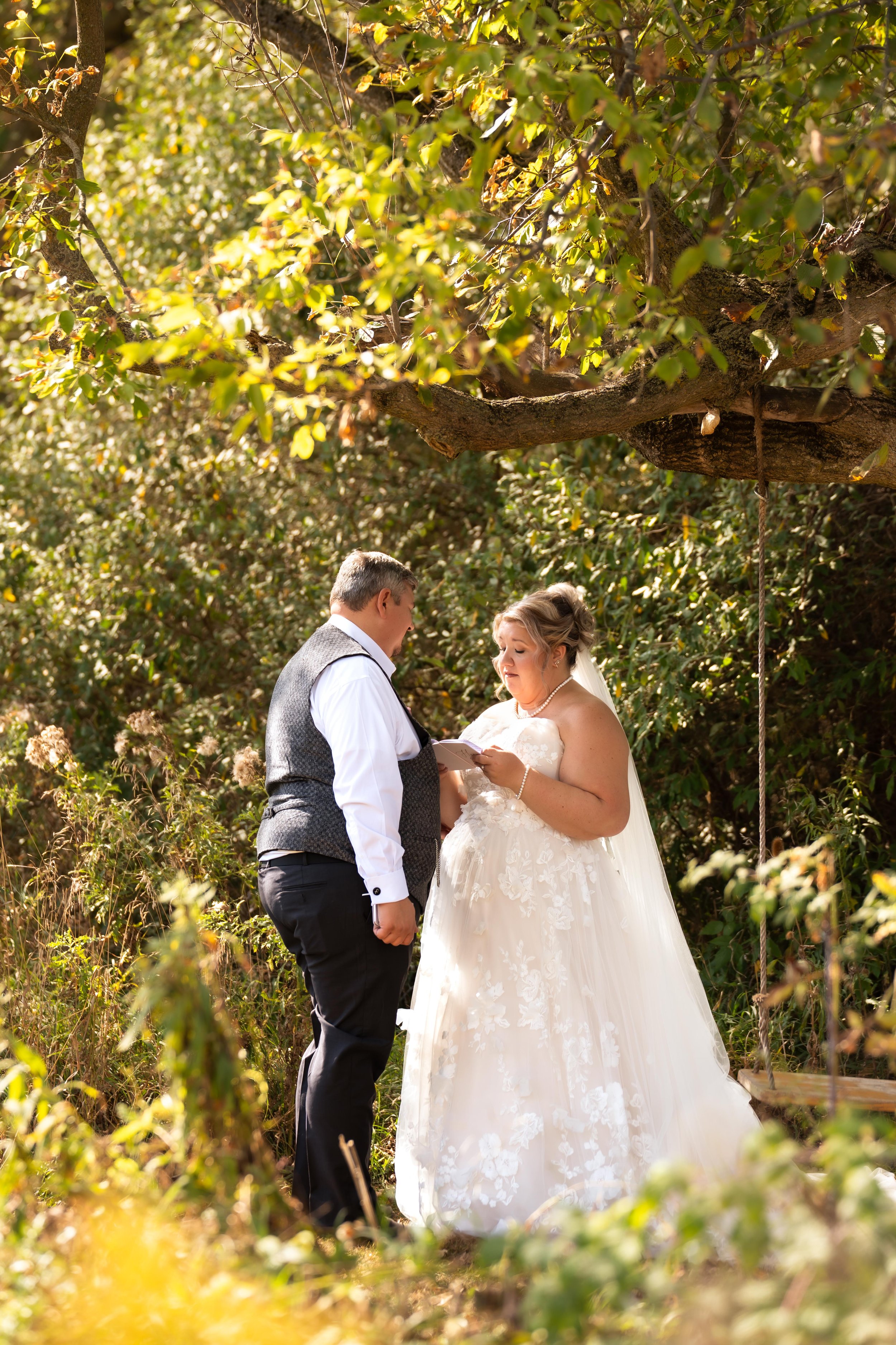 A bride and groom in wedding attire sharing an intimate moment outdoors under a tree, with the bride reading vows or a speech and the groom listening with a gentle expression.