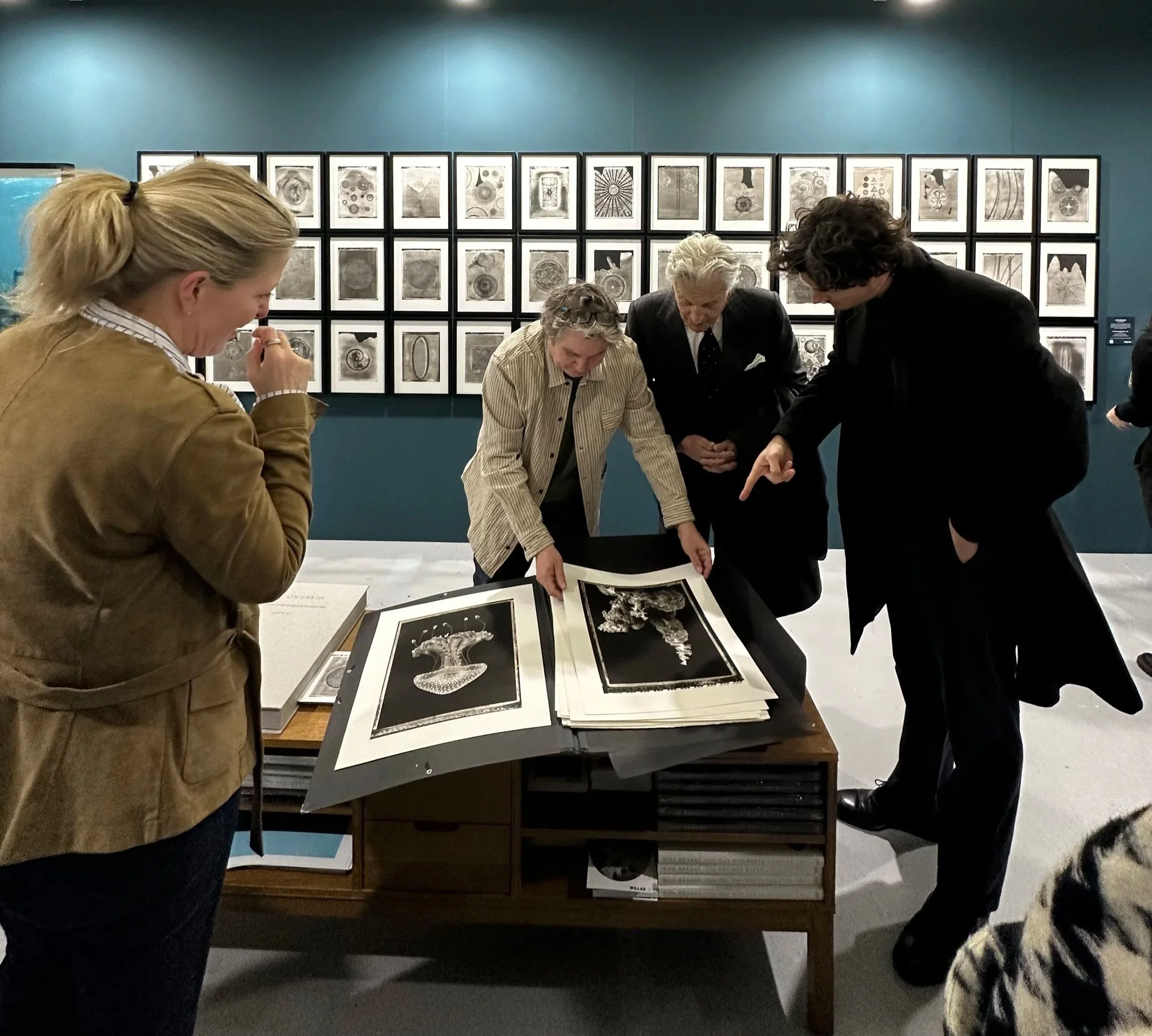 Four individuals examining black-and-white artwork displayed on a table at an art gallery, with framed art pieces on the wall behind them.