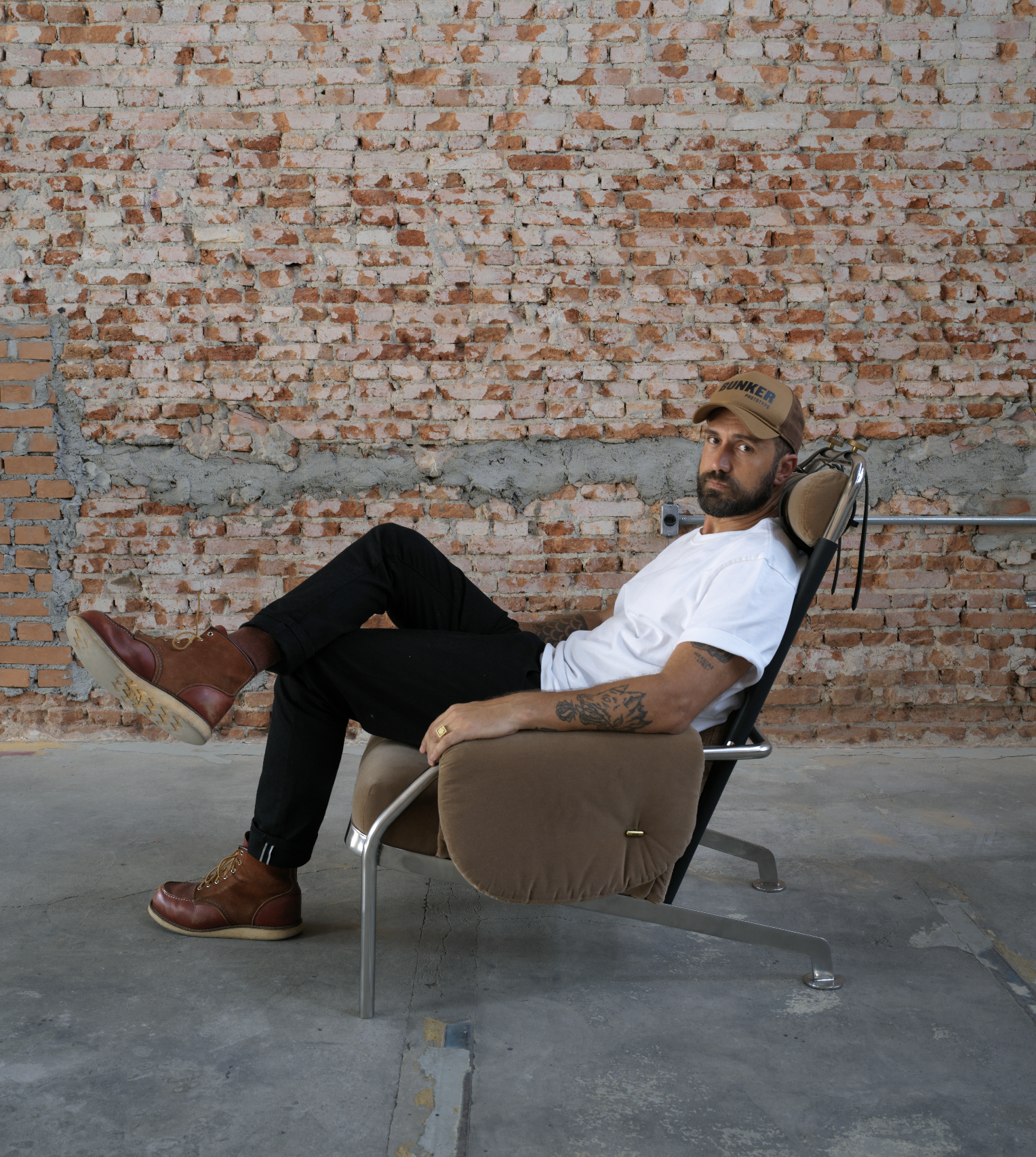 Homem com barba, tatuagens no braço, chapéu e camiseta branca, sentado em cadeira confortável com fundo de parede de tijolos expostos.