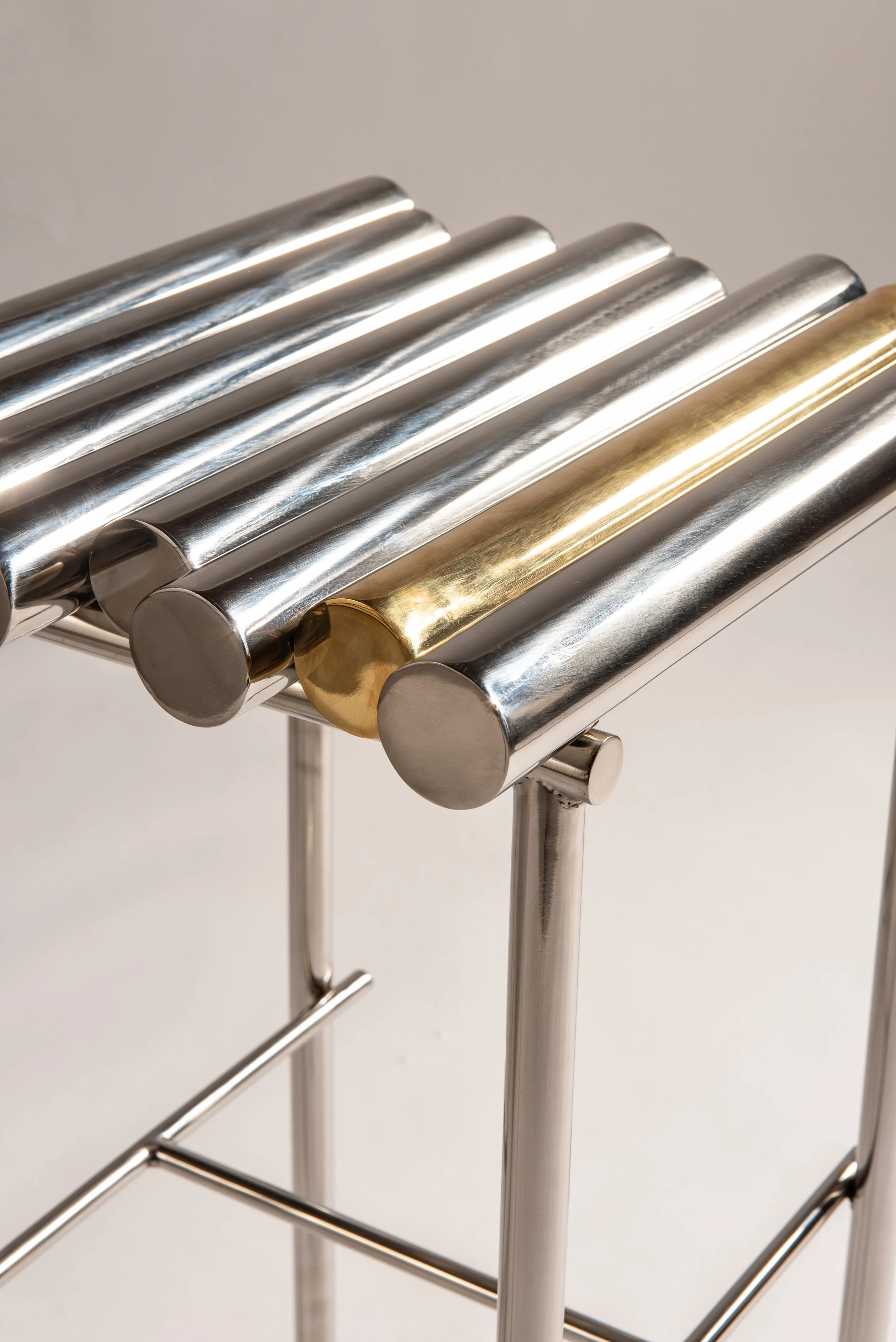 Banco de metal com tubos cromados e uma peça dourada, suportado por uma estrutura de suporte de aço.