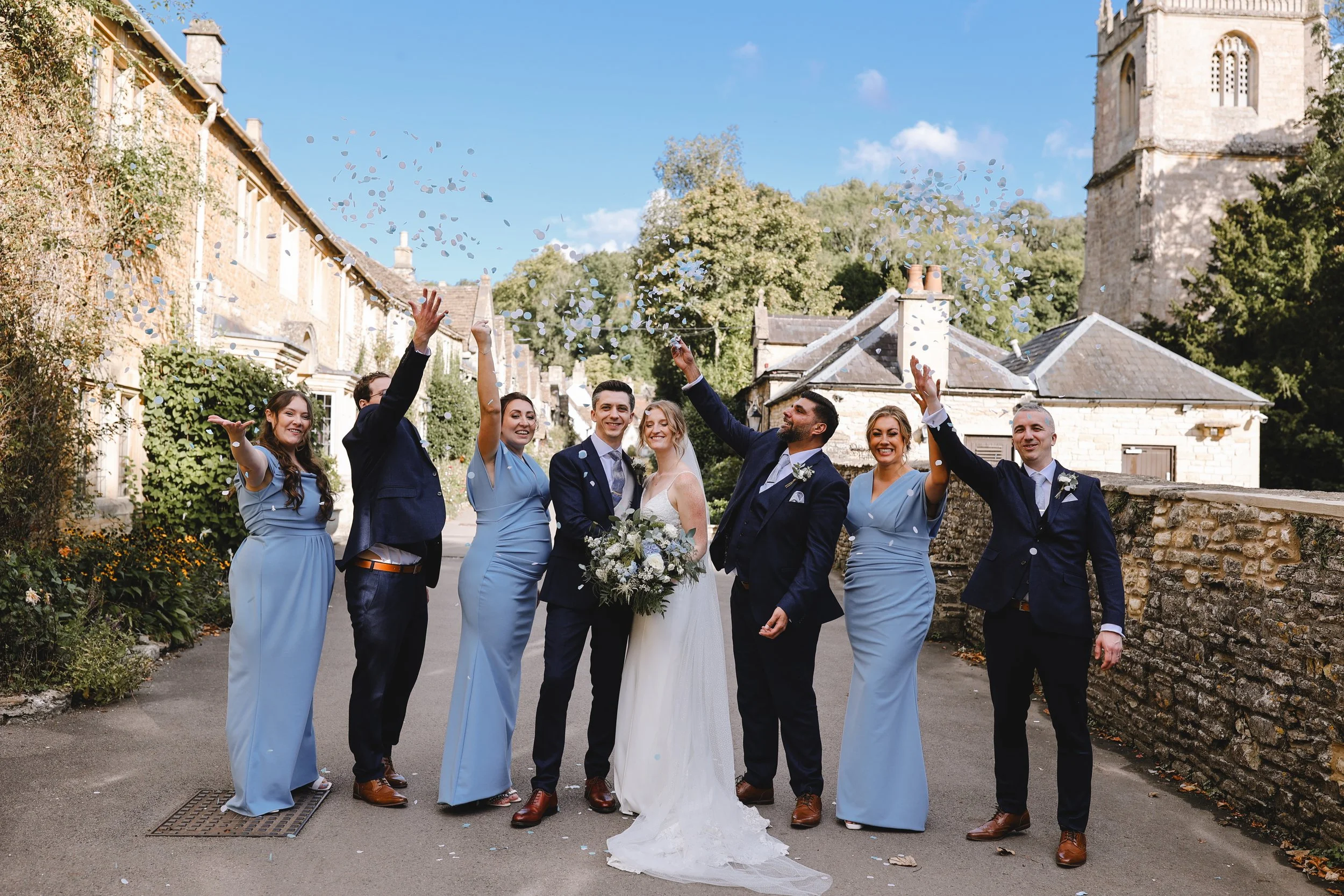 Wedding party celebrating outdoors with bride and groom holding a bouquet, surrounded by bridesmaids in blue dresses and groomsmen in suits, tossing confetti in a stone-walled village setting.