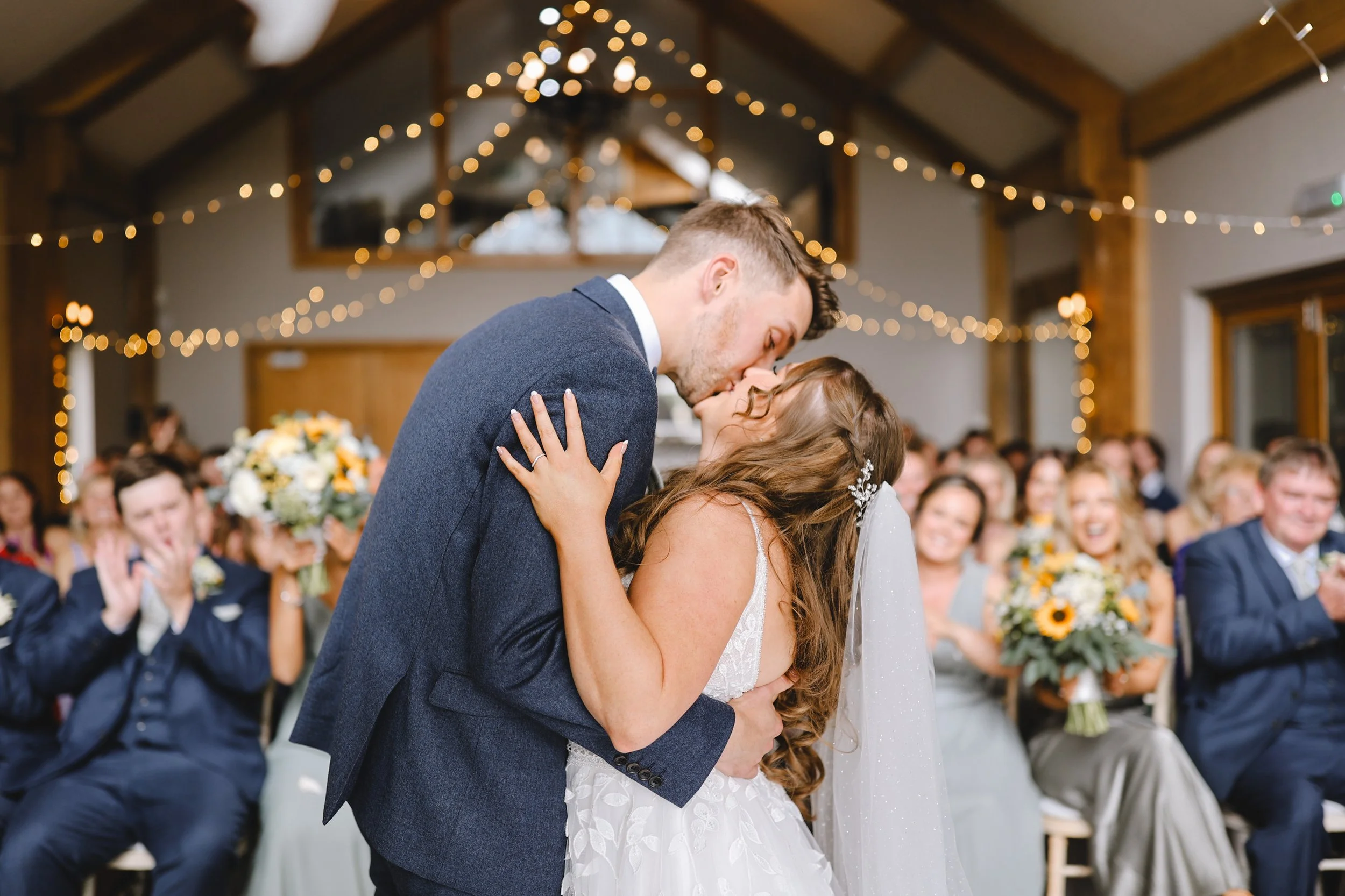 A bride and groom kissing at their wedding ceremony, surrounded by applauding guests, with soft string lights above.