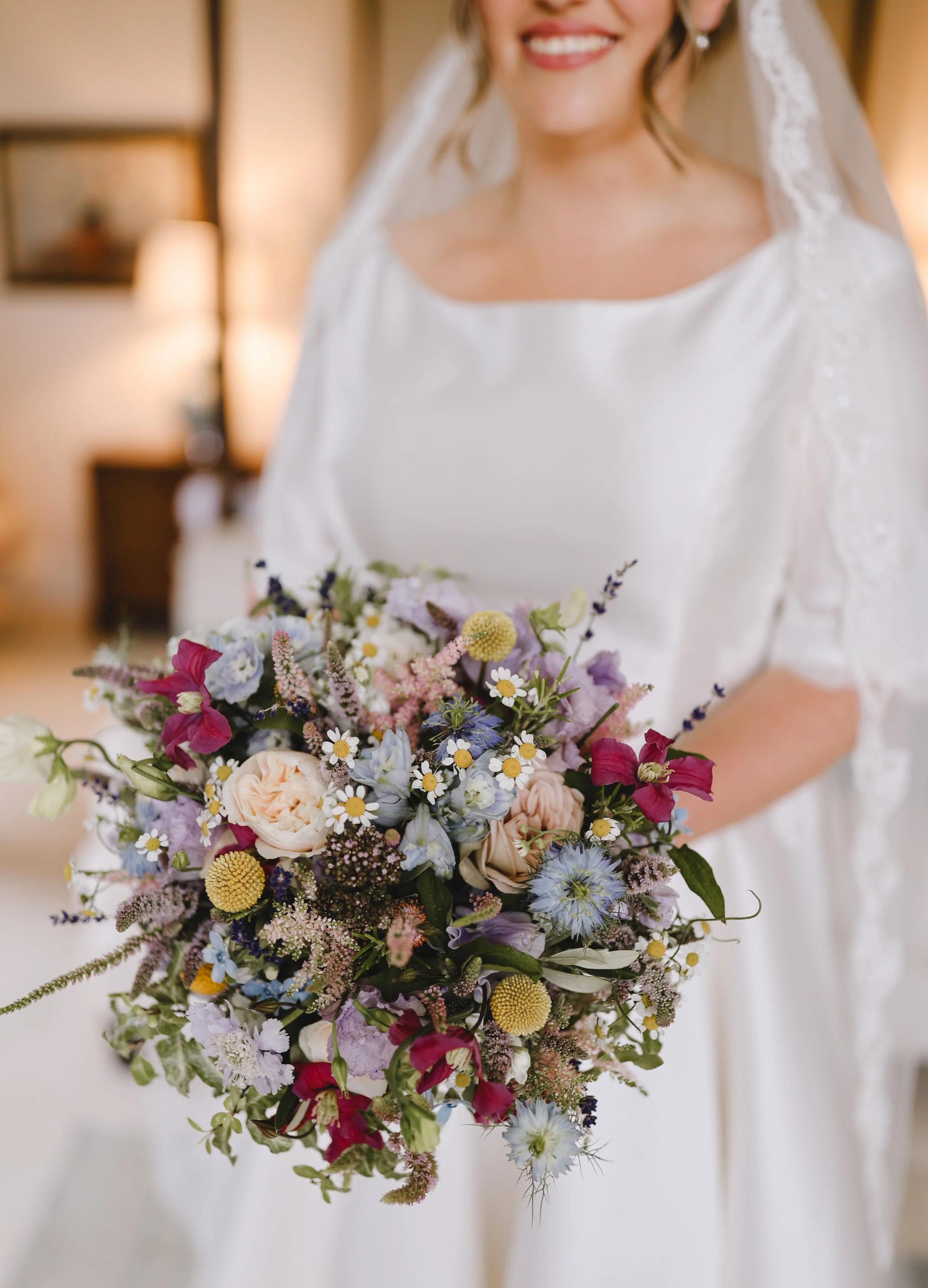 Bride holding a colorful bouquet with roses, daisies, and assorted wildflowers in a white dress with veil.