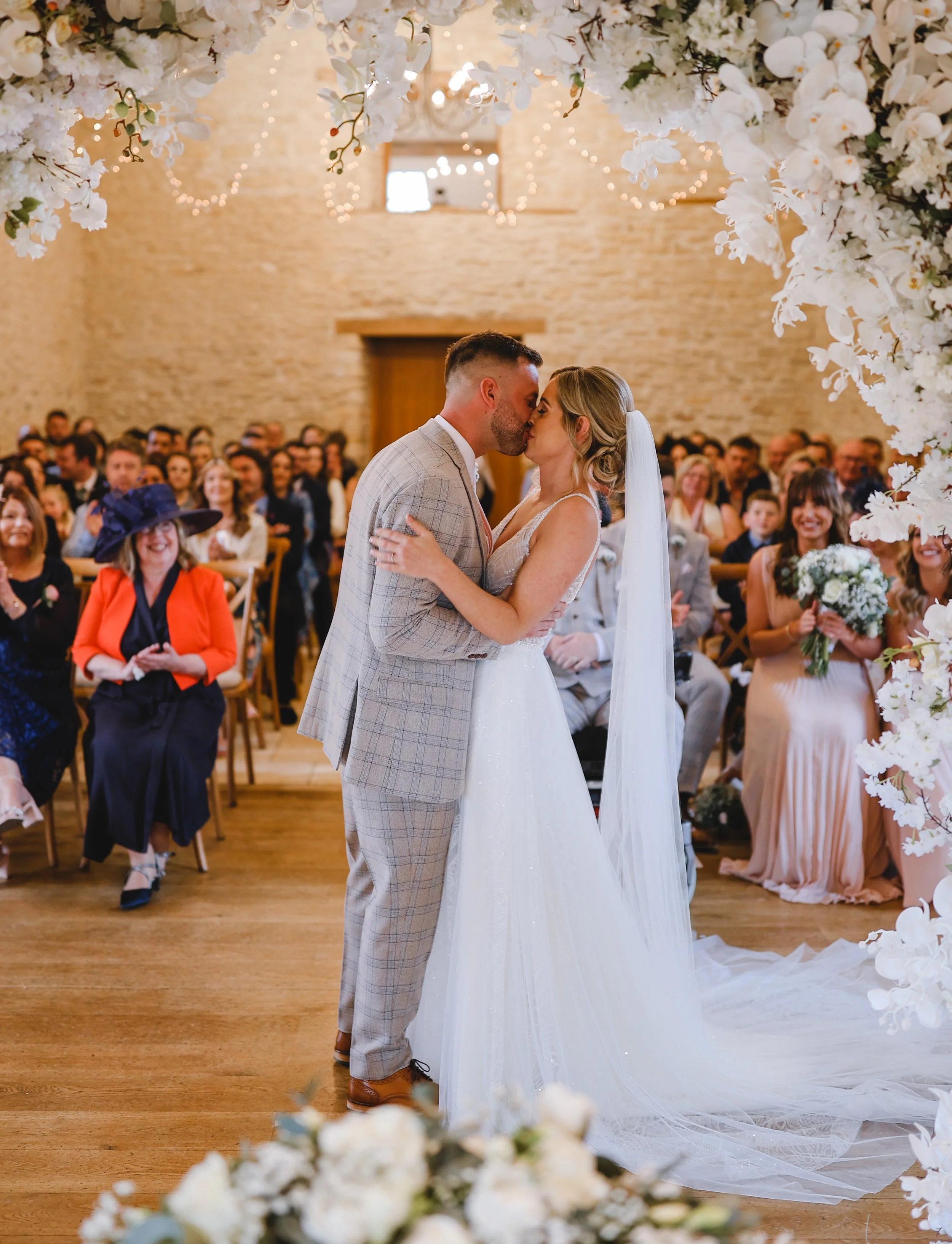 Bride and groom kissing at indoor wedding ceremony with floral decorations and guests seated.