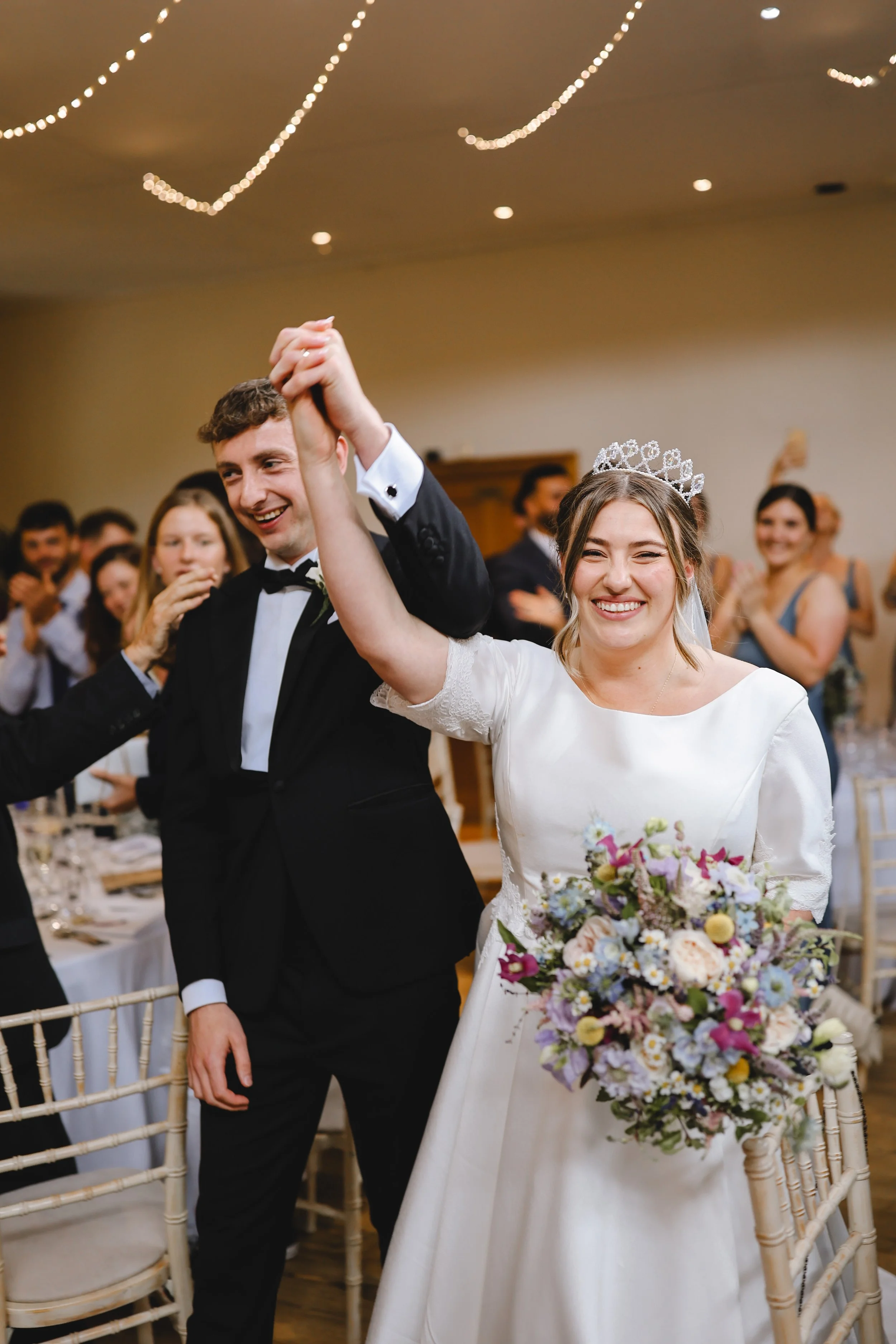 A bride and groom celebrating at a wedding reception, smiling and holding hands above their heads. The bride is wearing a white dress and tiara, holding a colorful bouquet. The groom is in a black suit. Guests are clapping in the background.