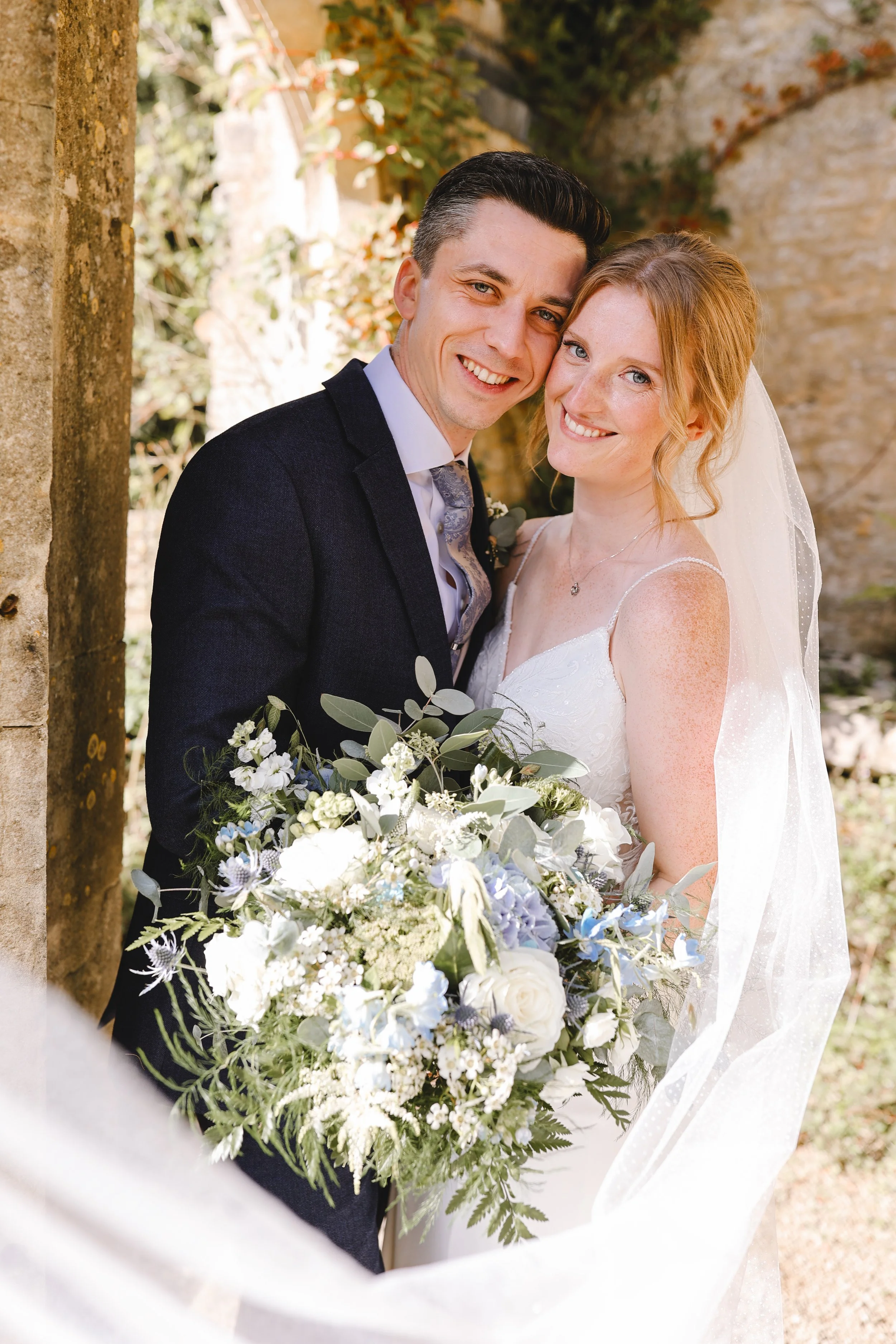 Bride and groom posing outdoors with a large floral bouquet and stone background