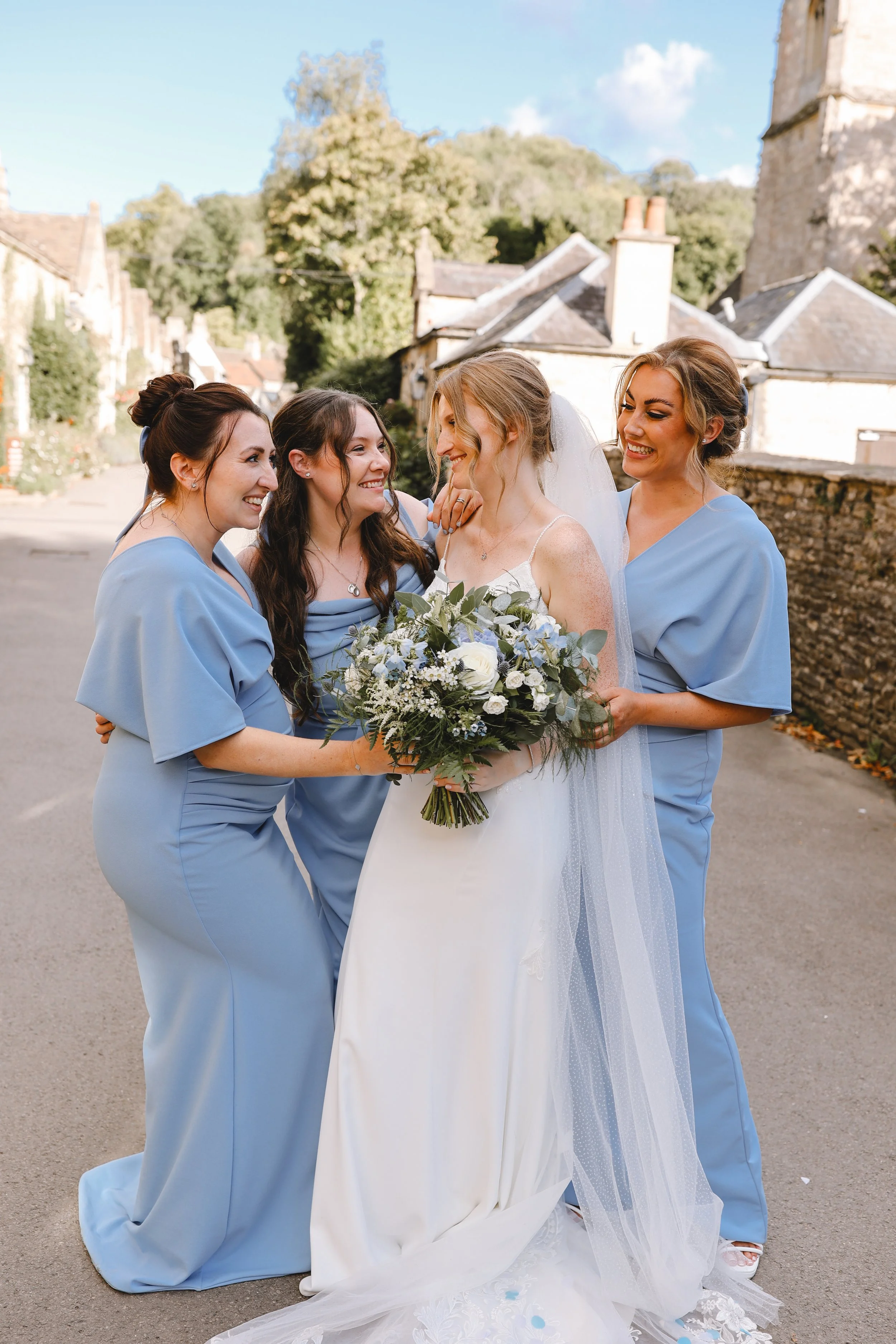 Bride in white dress with three bridesmaids in light blue dresses, all smiling and standing outdoors on a sunny day.
